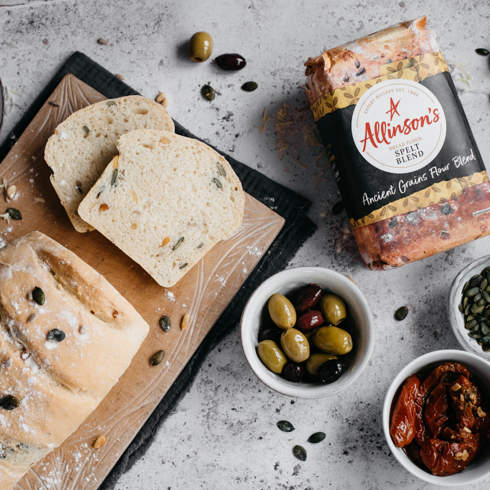 Overhead shot of spelt loaf with two slices in front, bread flour, olives and tomatoes