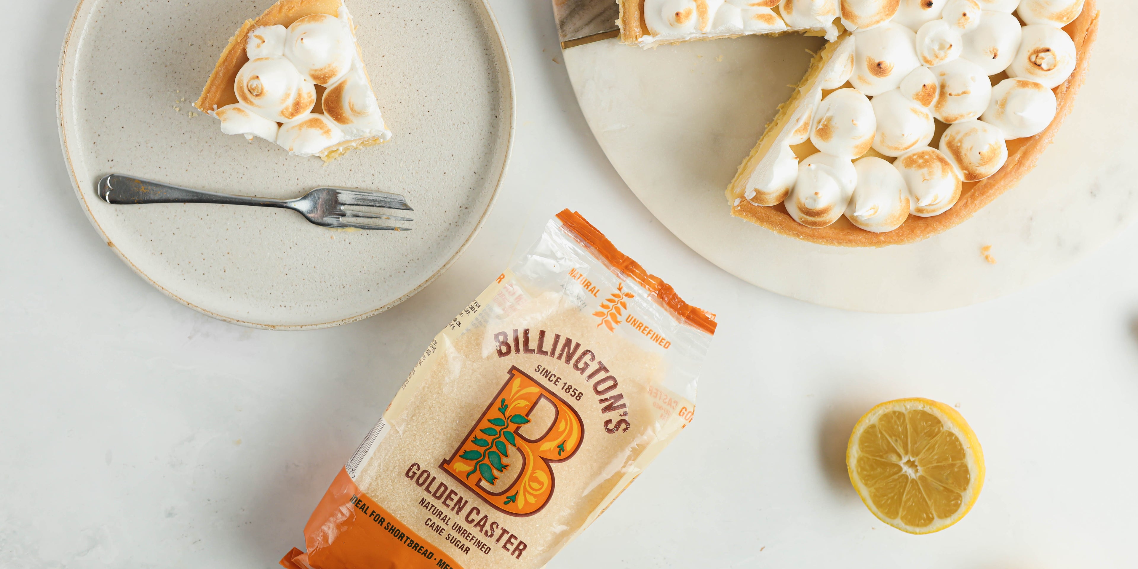 A slice of lemon meringue pie on a white plate with a fork. Next to it a bag of Billington's sugar and the remaining lemon meringue pie on a white plate