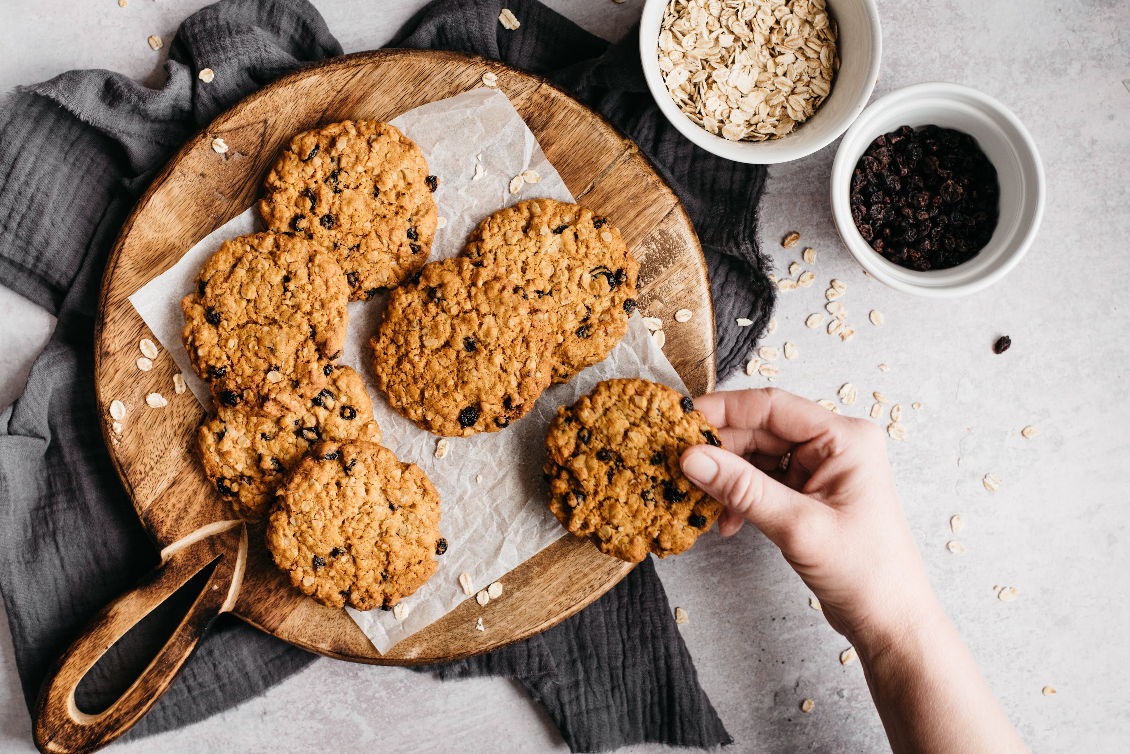 Top view of a batch of Oat & Raisin Cookies on baking parchment ready to serve, with a hand reaching for a cookie