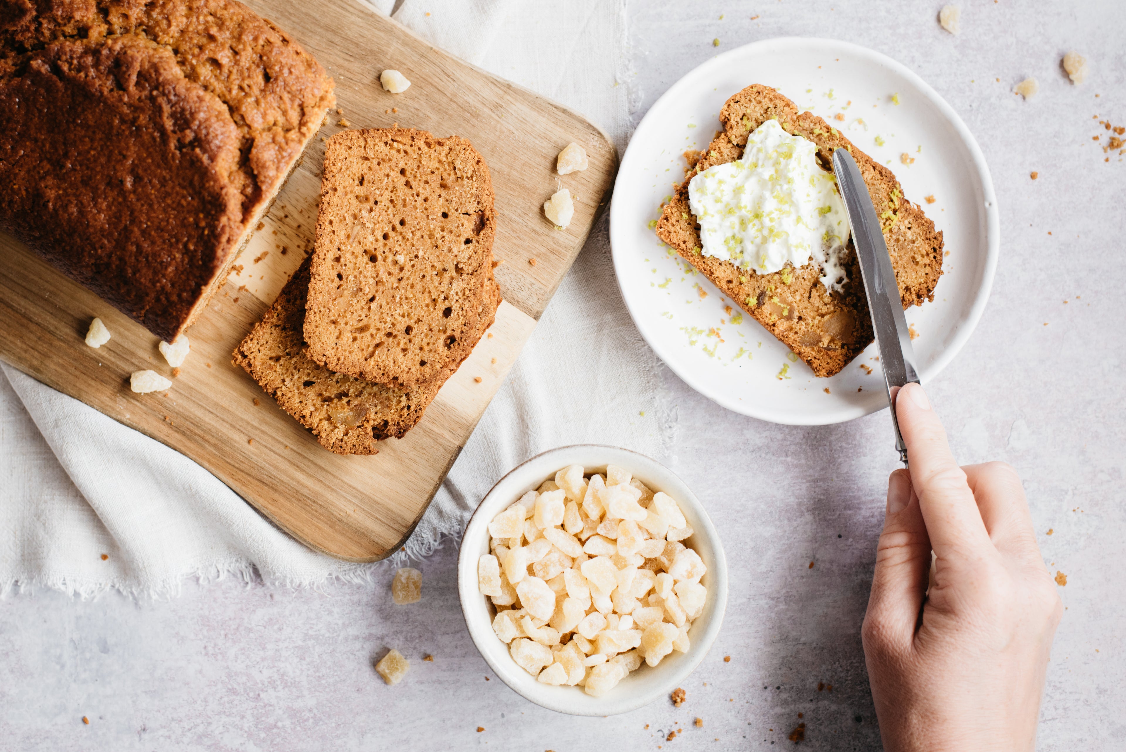 Flat lay of Gingerbread Loaf on a serving board, next to a bowl of ginger and a plate with a slice of Gingerbread Loaf with a hand buttering it