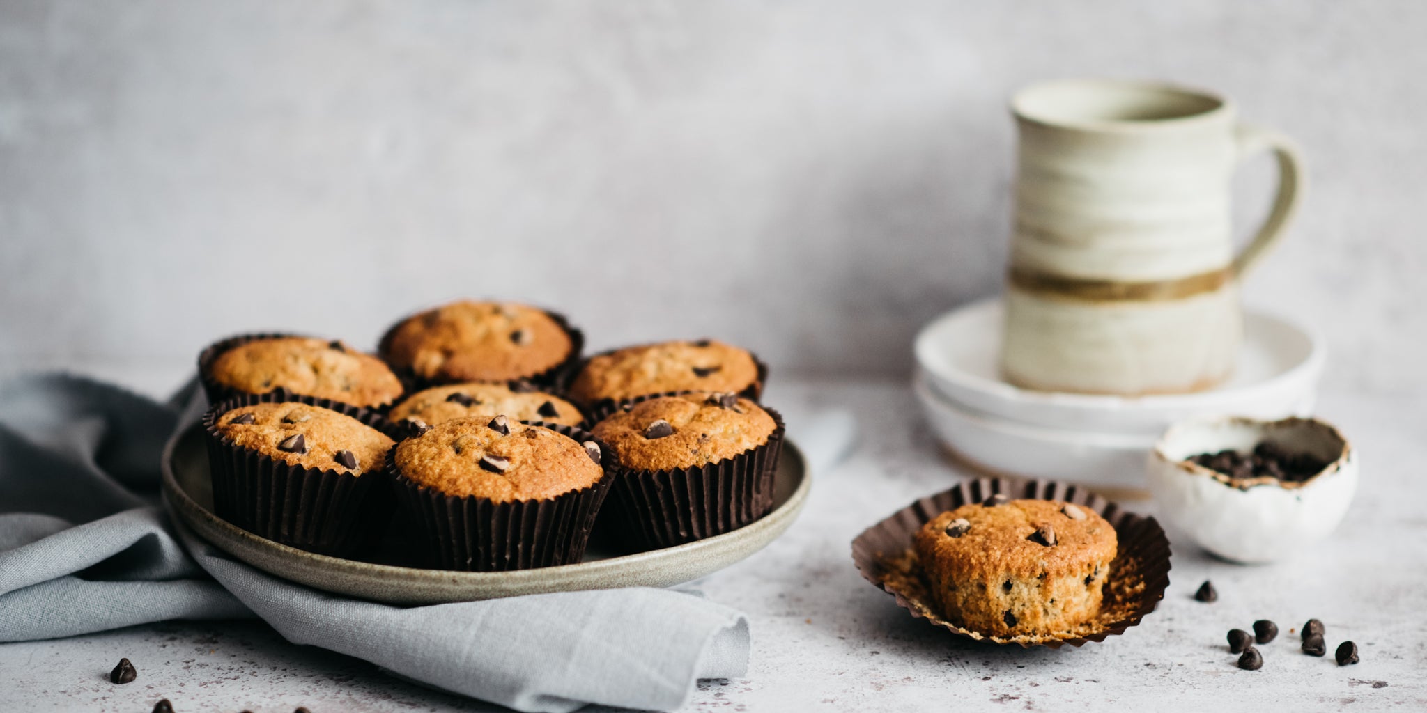 Wholemeal Banana & Chocolate Chip Muffins on a serving plate next to a chocolate chip muffin with it's case peeled back ready to eat.