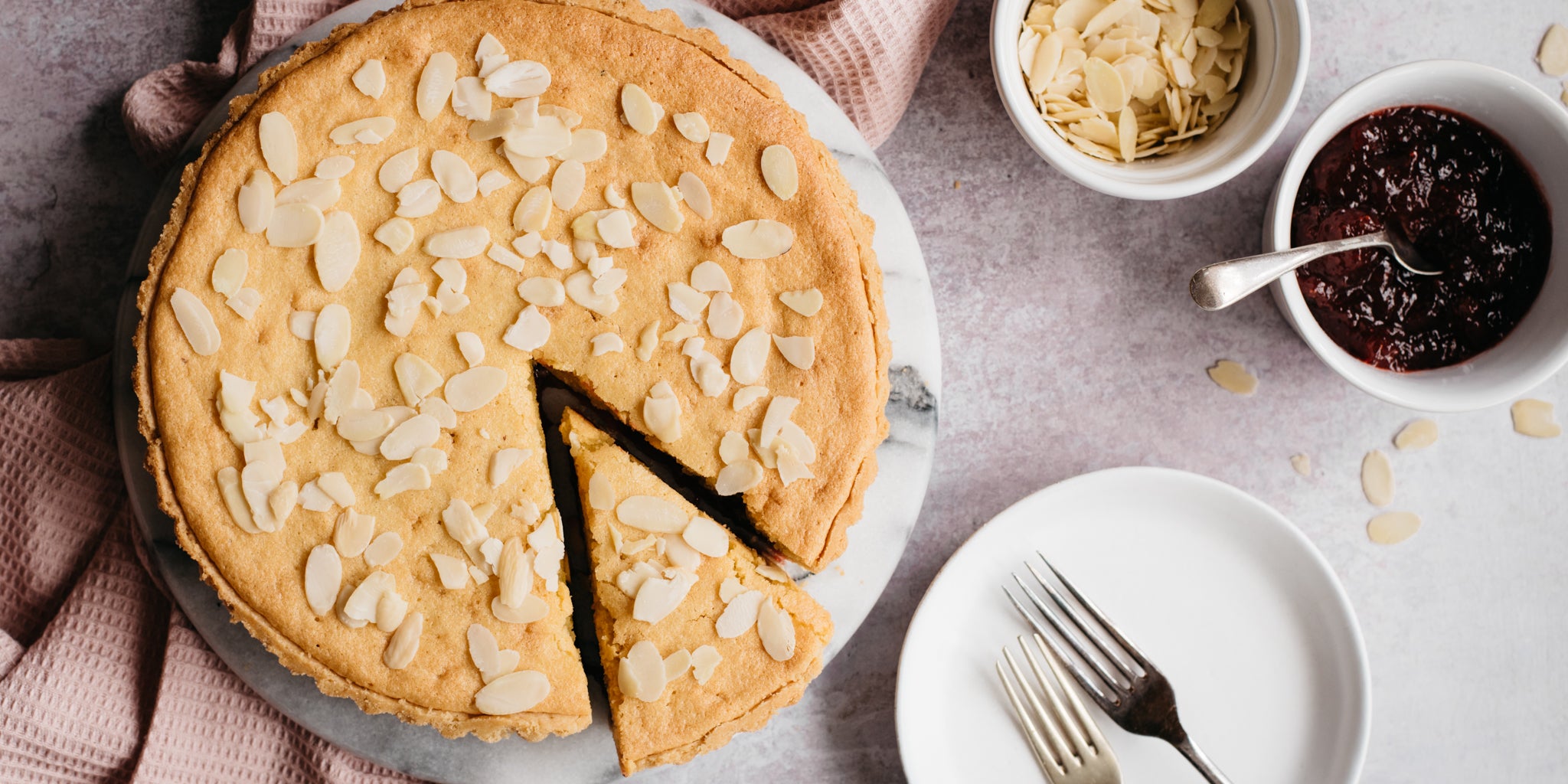 Simple Bakewell Tart with a slice cut out of it ready to serve, next to plates, forks and a bowl of jam from the filling