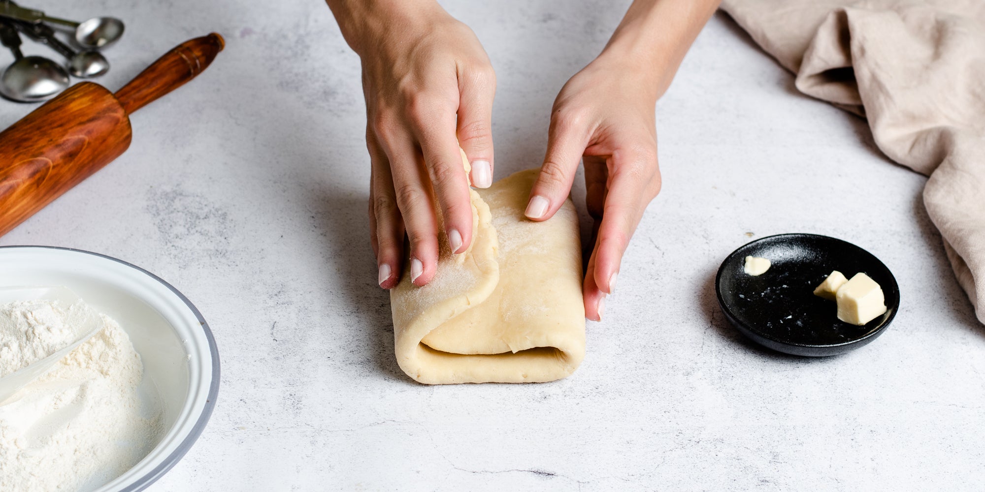Puff Pastry being hand folded by hands, next to a dish of butter and a bowl of flour and a rolling pin