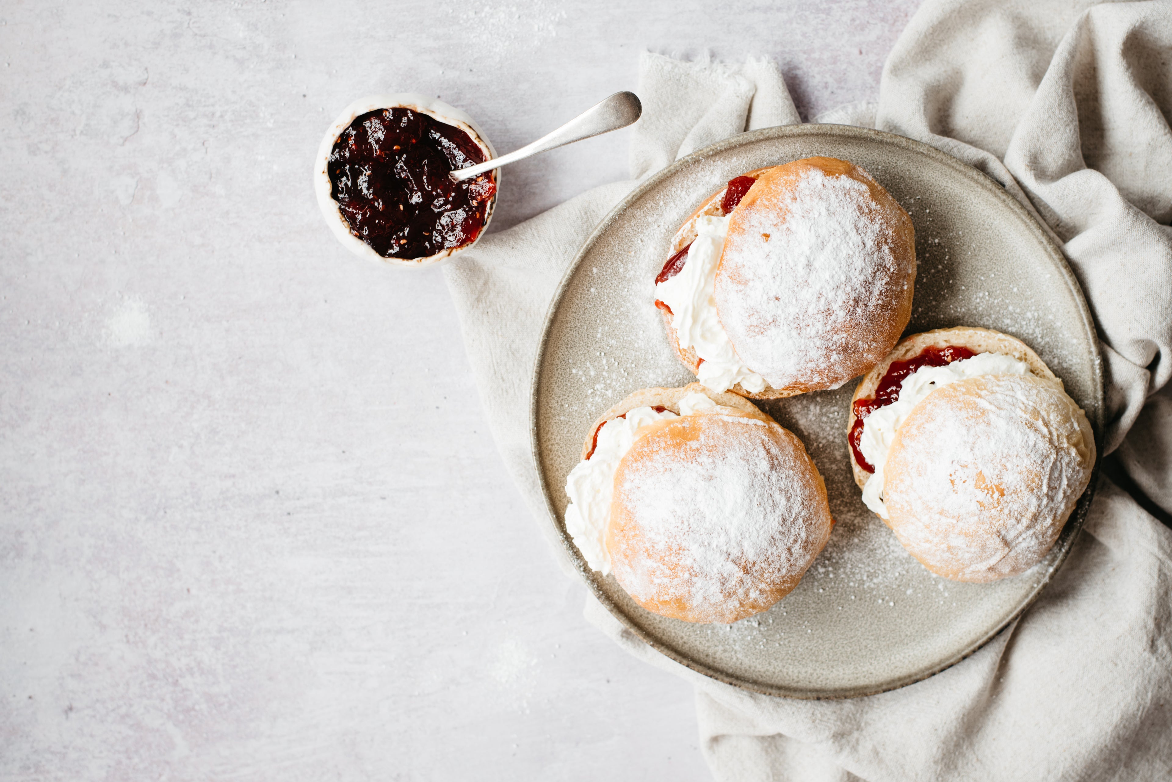 Top view of Devonshire Splits on a plate, dusted with icing sugar next to a bowl of jam with a spoon
