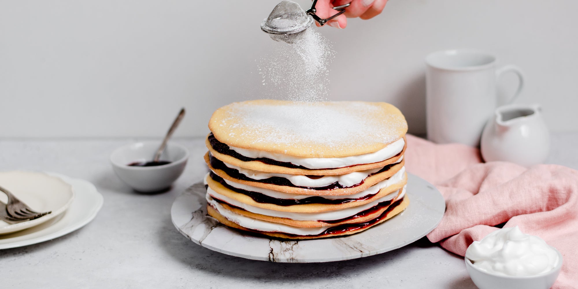 Danish Layer Cake being dusted with icing sugar by a hand, on a marble serving plate, a bowl of whipped cream in the foreground