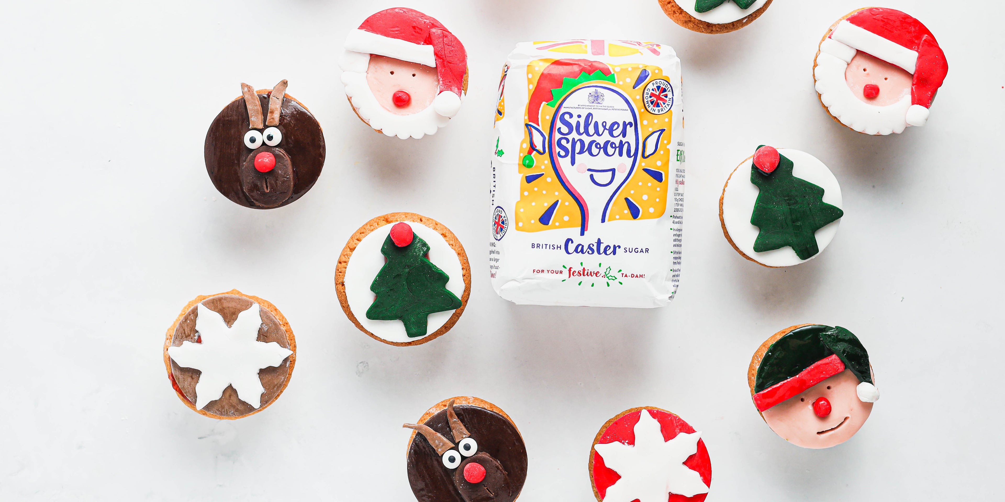 Top view of Christmas Cupcakes with reindeer, Christmas tree and santa fondant decorations, next to a bag of silver spoon caster sugar