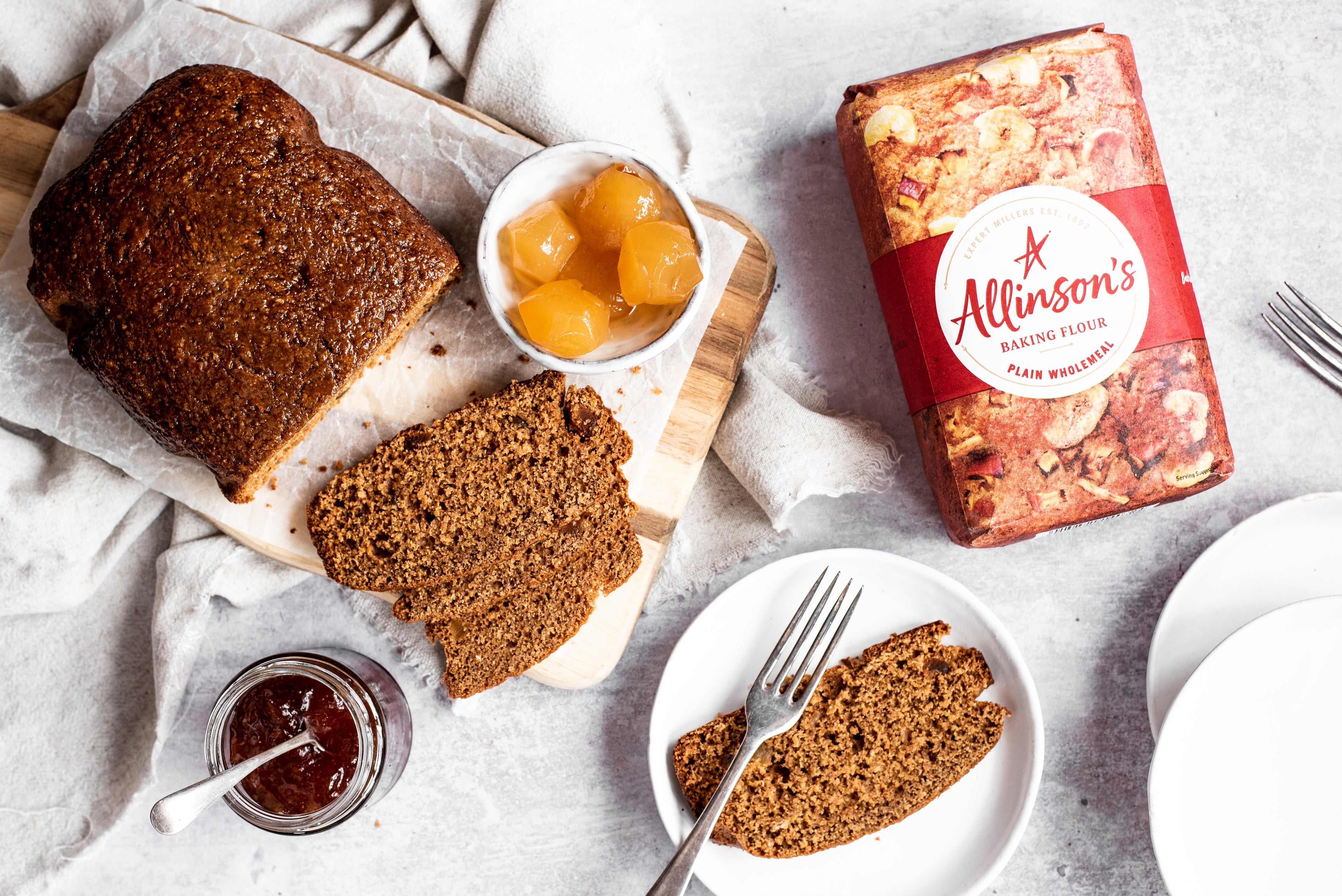 Overhead shot of gingerbread loaf with slices cut in front. Slice of cake on plate with fork. Bowl of ginger. Marmalade jar. Flour pack