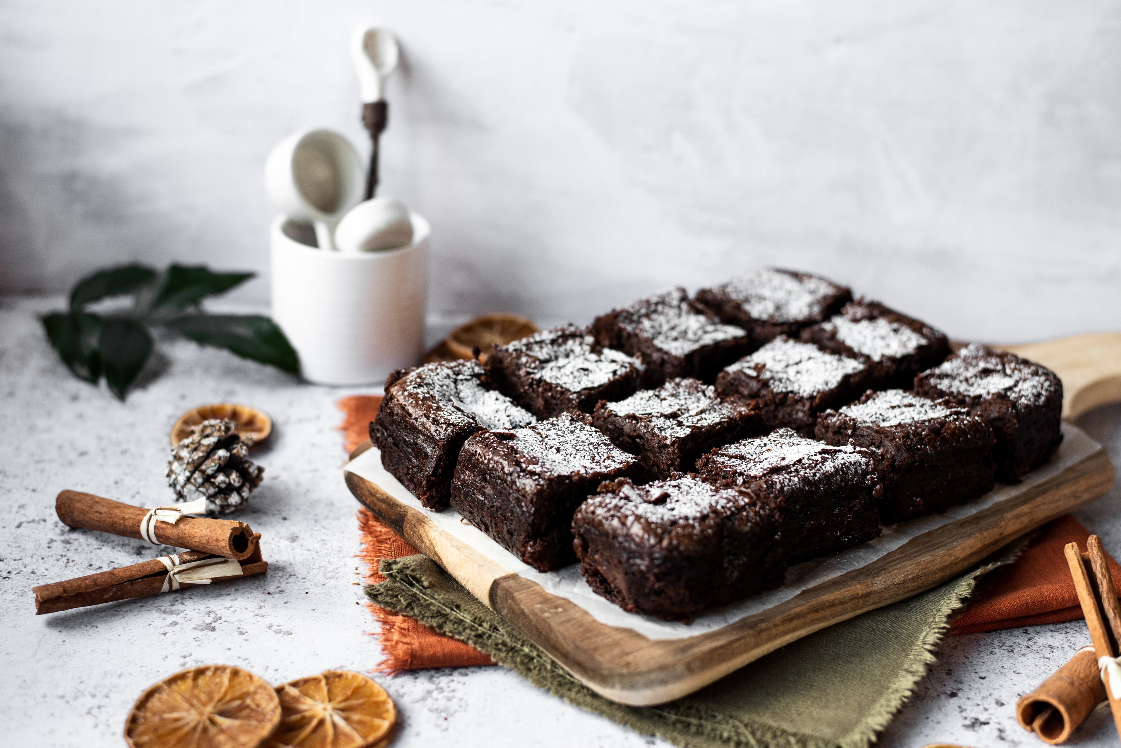 Full shot of brownie chopped into squares on chopping board with cinnamon sticks and dried orange. 