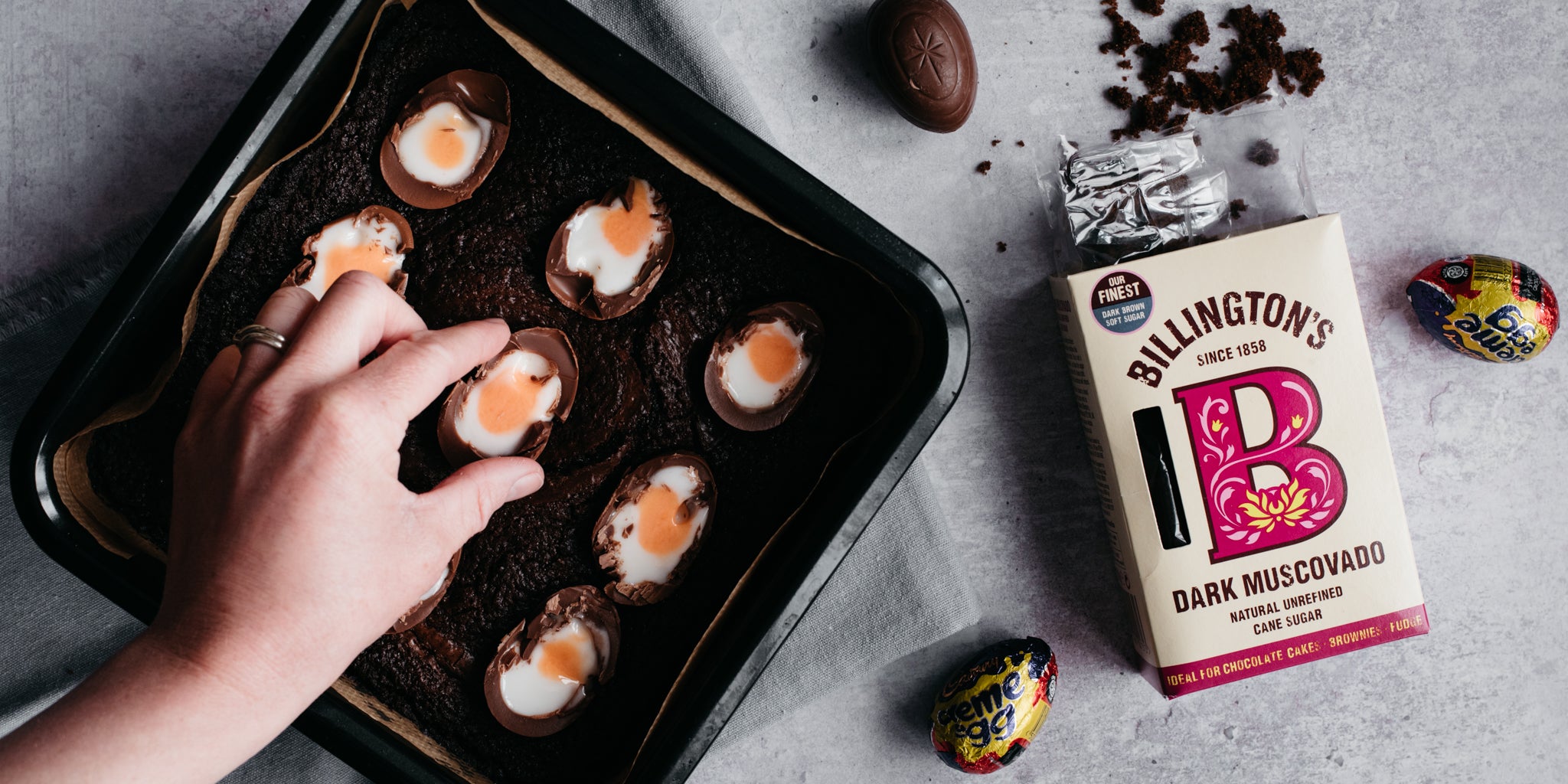 Hand placing creme eggs in the centre of brownie mixture with open pack of sugar and creme eggs beside