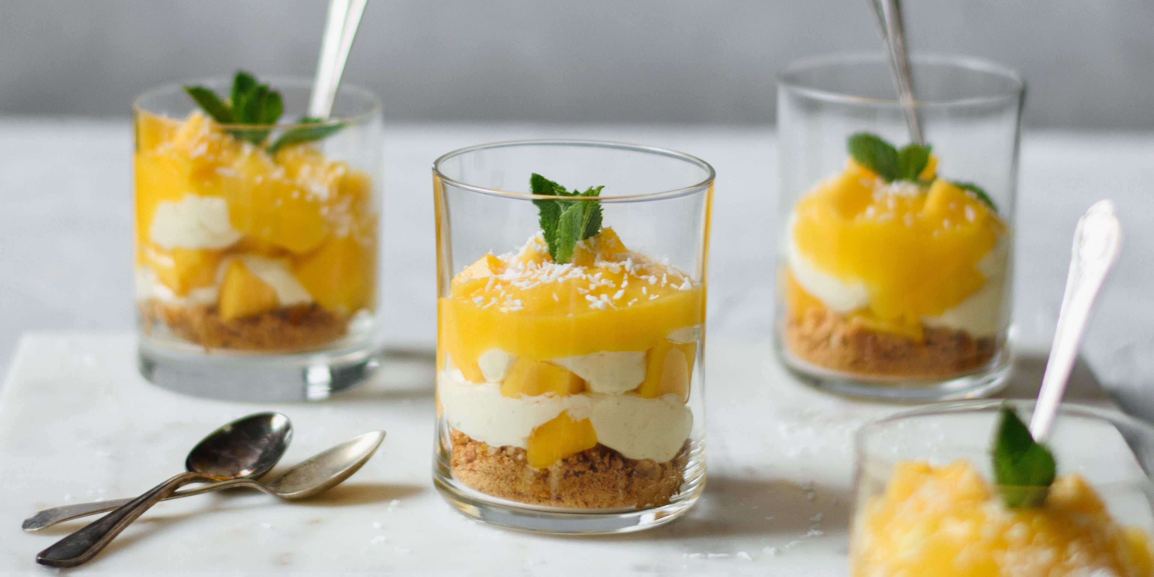 3 small glasses of mango syllabub with spoons in and sprig of mint on top. Two spoons on the side