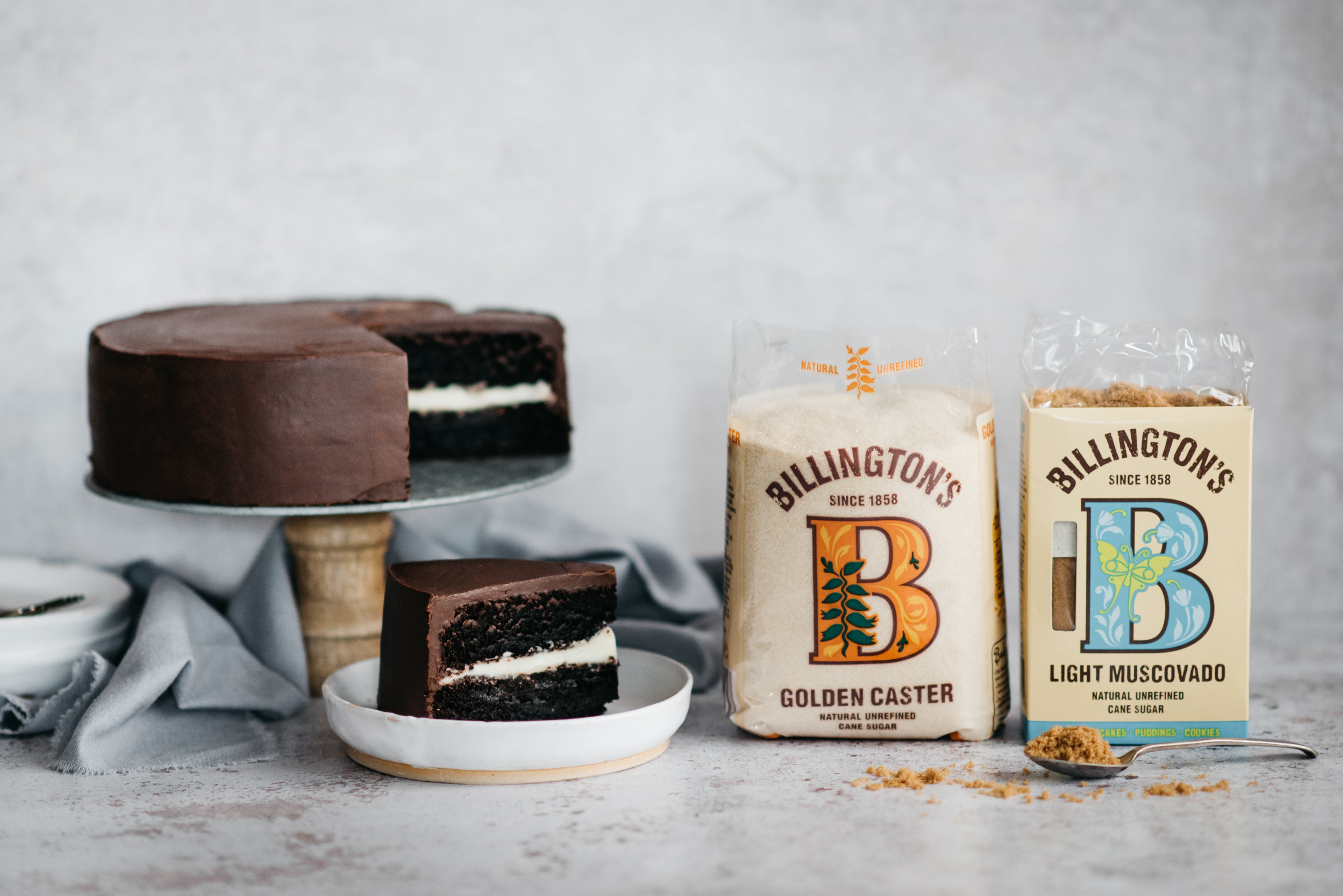 Best Chocolate Cake by Billington's on a cake stand, with a slice cut out showing the cream layer inside. Next to a bag of Billington's Golden Caster and Light Muscovado Sugar