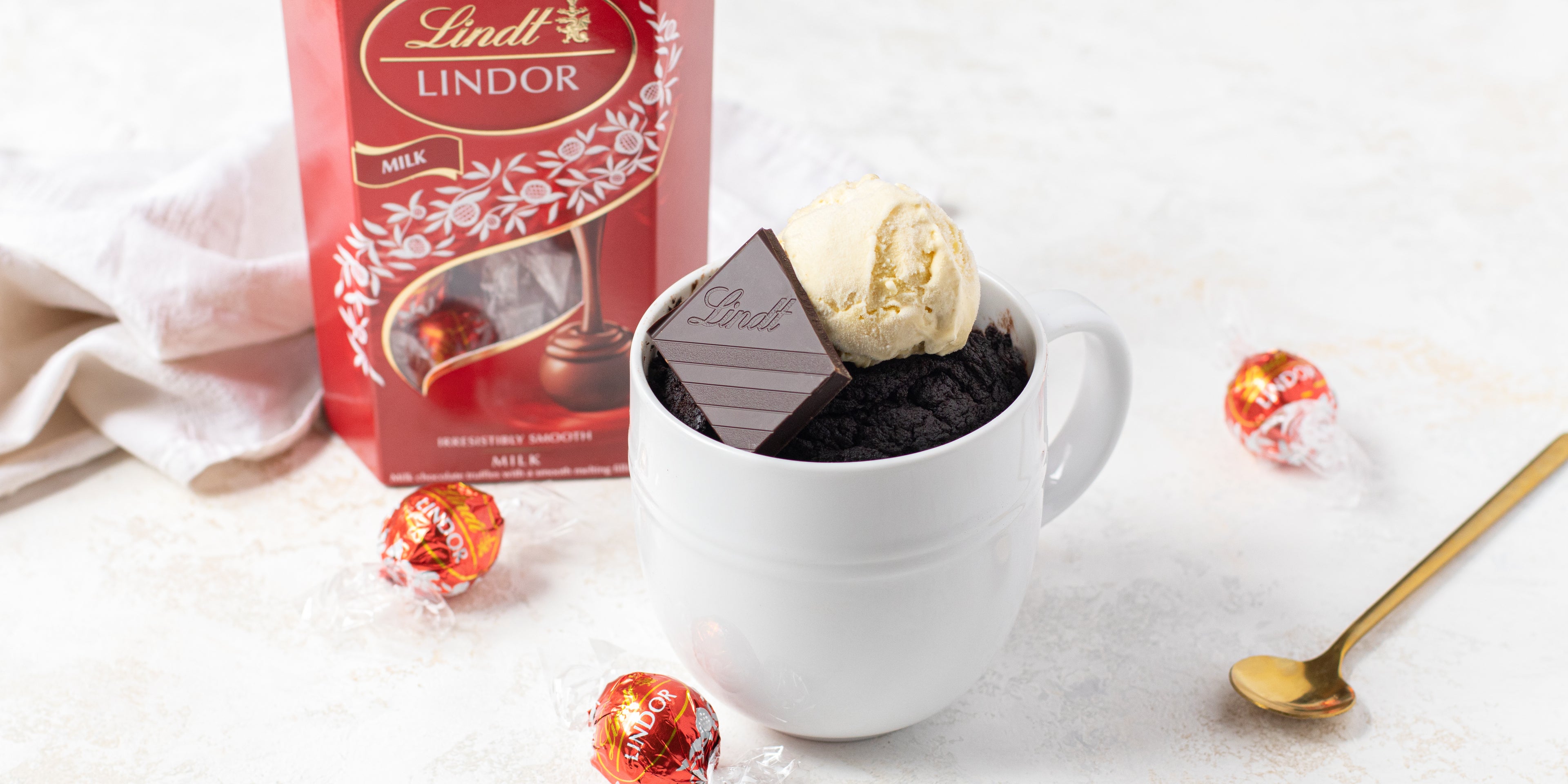 Chocolate Mug Cake customised with Lindt chocolate and a dollop of vanilla ice cream. In the background these is a box of Lindt balls