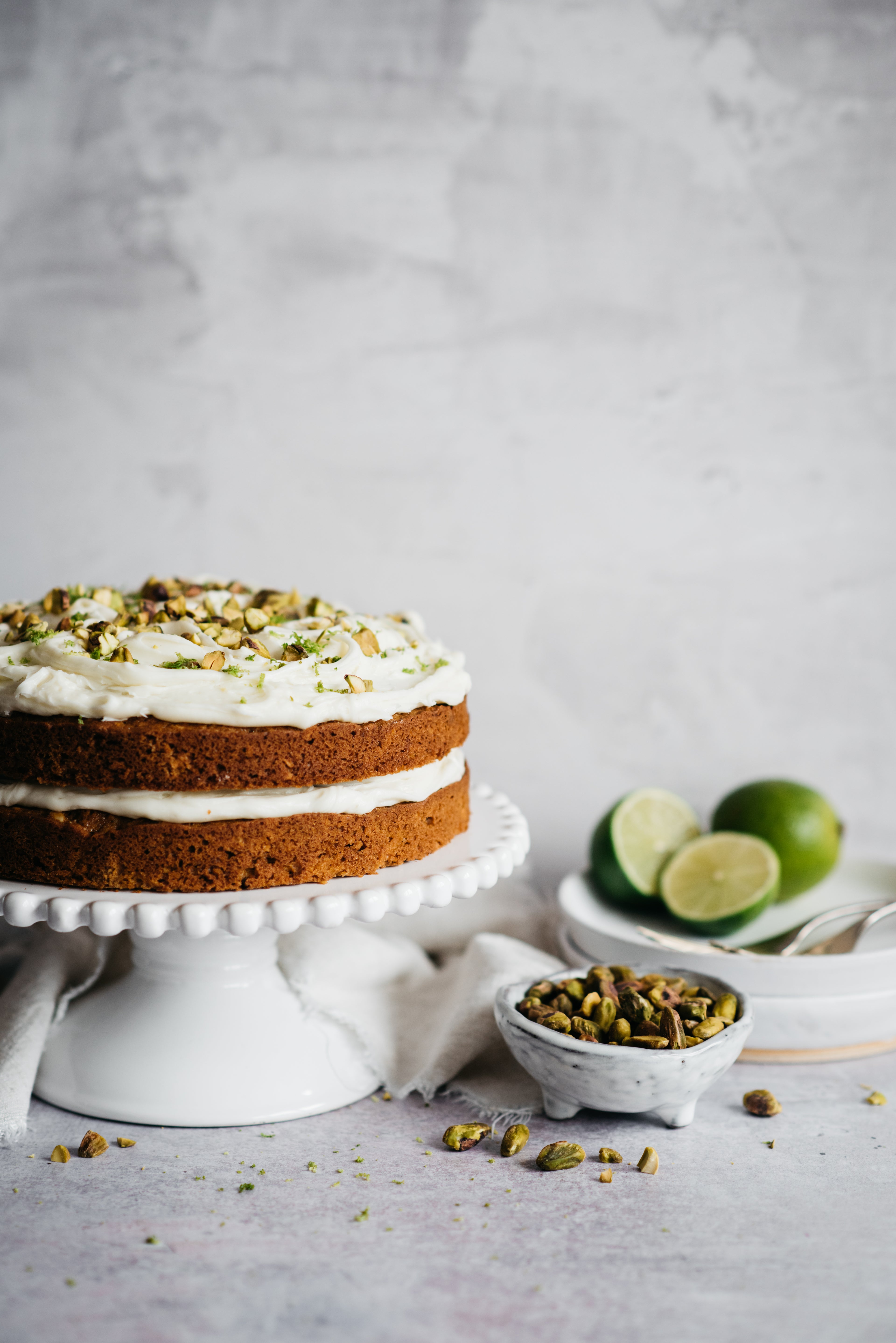 Carrot cake with icing on cake stand. Bowl of pistachios and limes in foreground