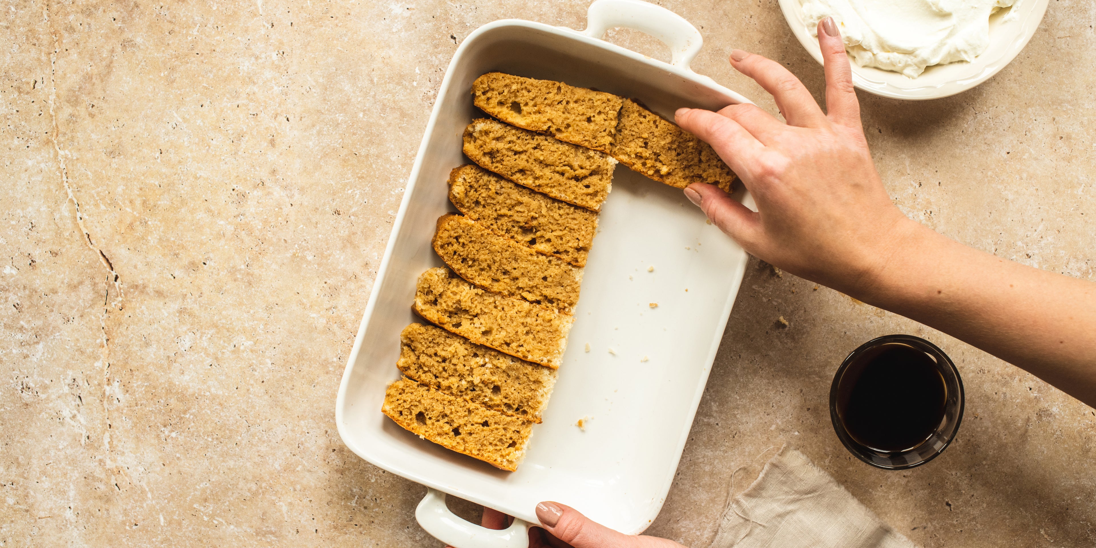 Vegan Tiramisu being prepared with hands lining the dish with coffee soaked biscuits, next to a cup of coffee and whipped vegan cream