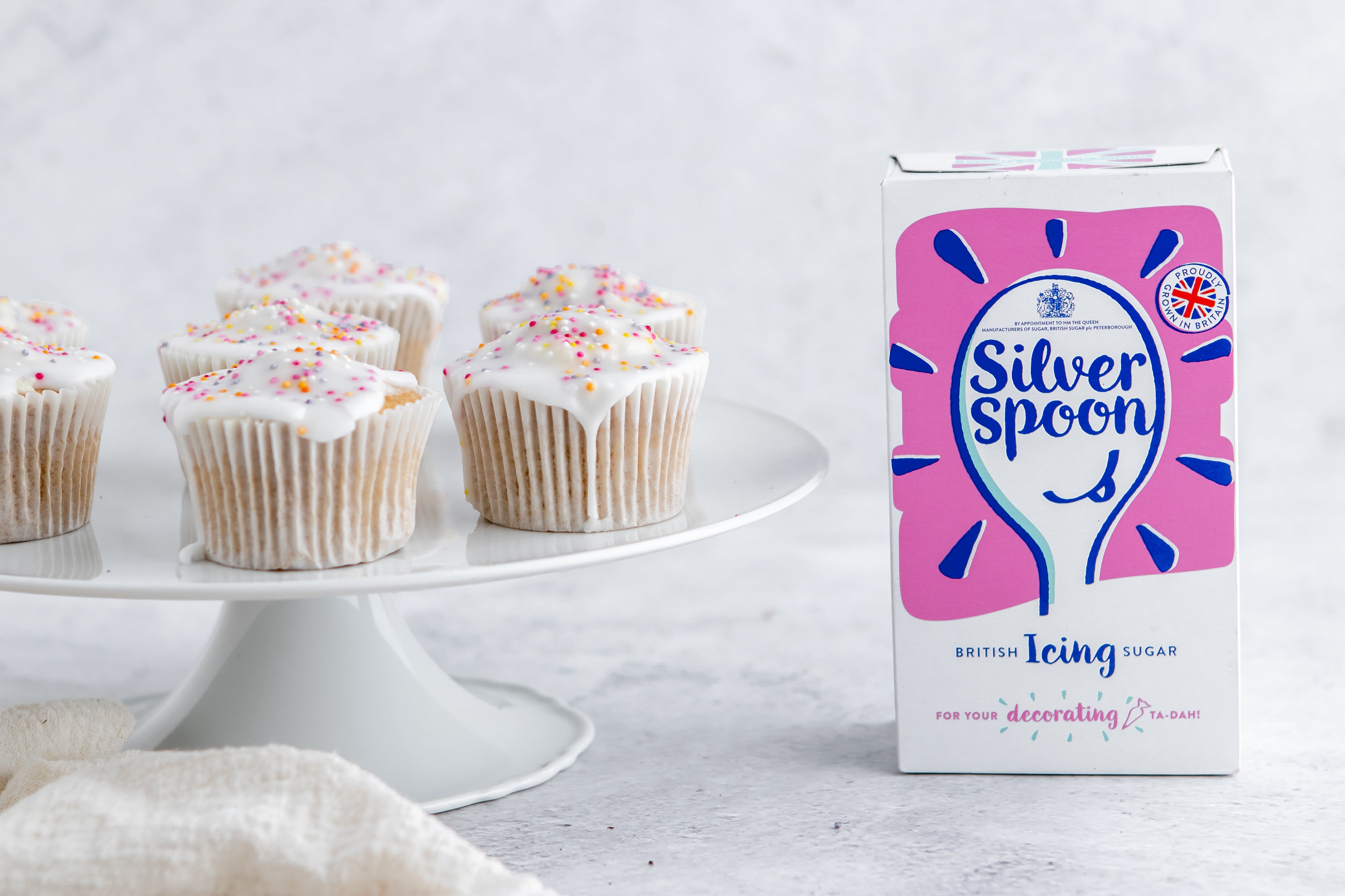 Simple Fairy Cakes on a cake stand, with sprinkles and running icing. Next to a box of Silver Spoon Icing sugar