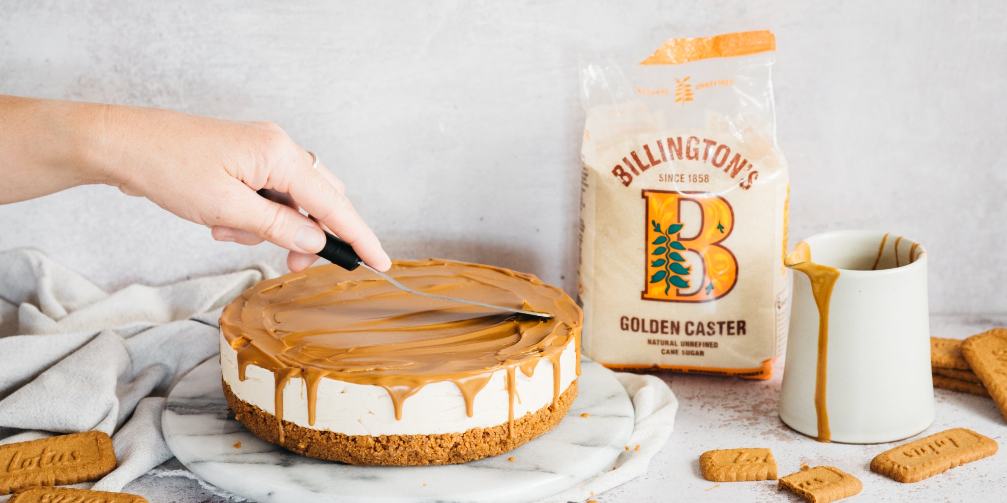 Cheesecake with hand spreading biscoff topping, beside a big bag of sugar