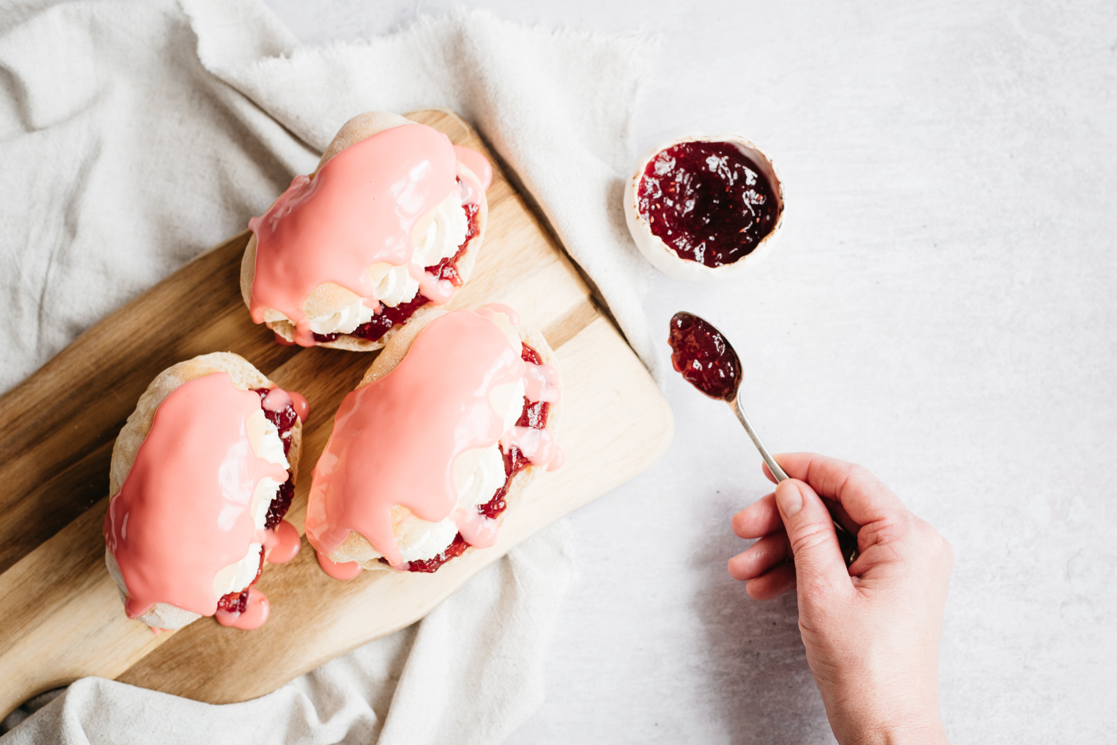 Top view of Iced Finger Buns on a serving board, next to a bowl of jam with a hand spooning jam onto the Iced Finger Buns