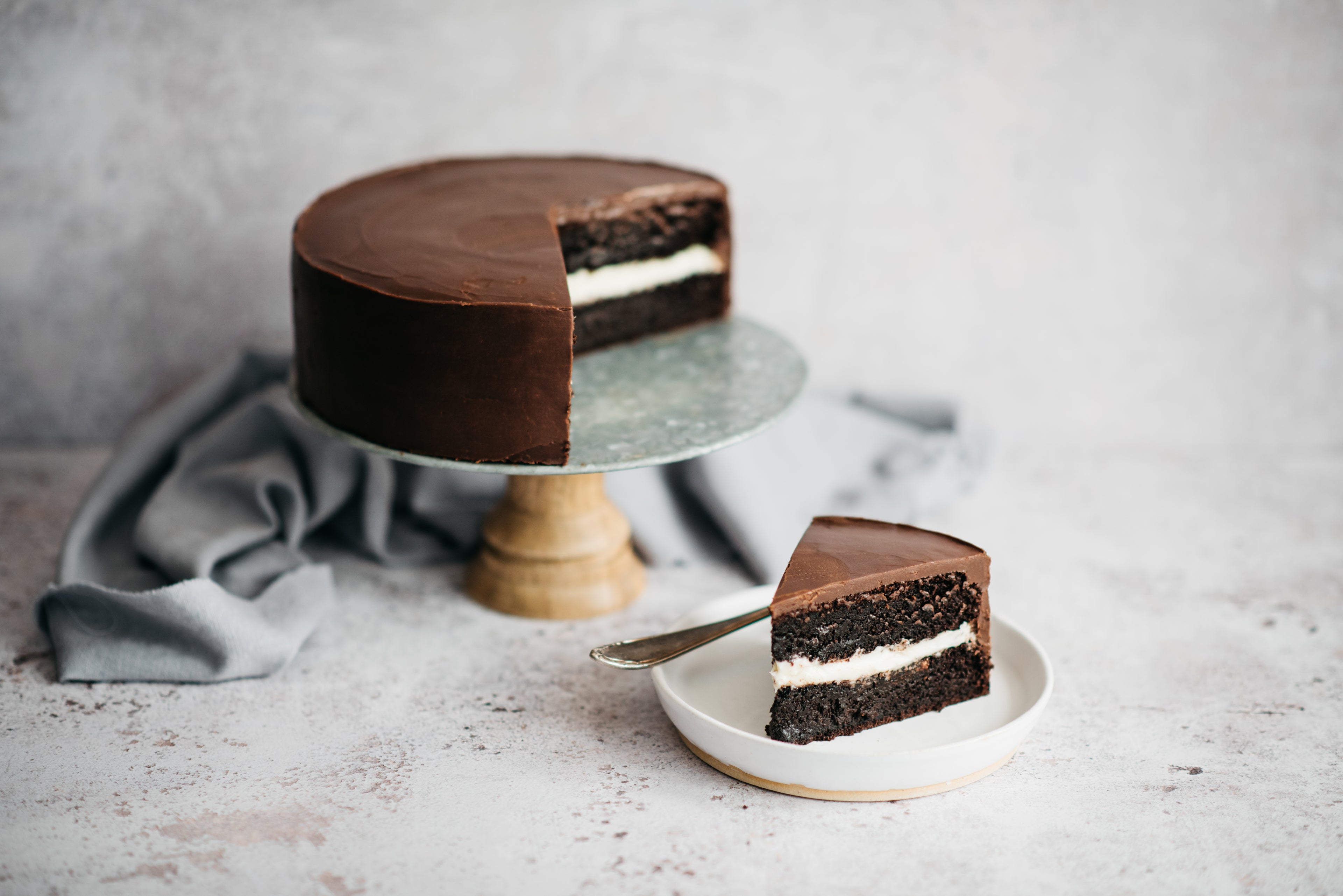 Best Chocolate Cake on a cake stand smothered in chocolate ganache with a slice cut out and served on a plate showing the creamy layer inside