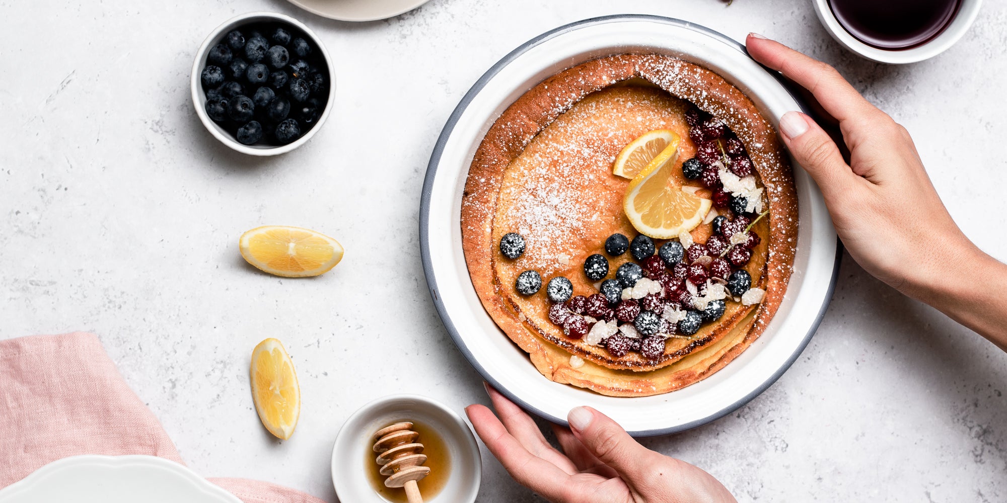 A plate of Dutch Baby Pancakes with hands, topped with berries and lemon, next to a honey drizzler and a small bowl of blueberries 