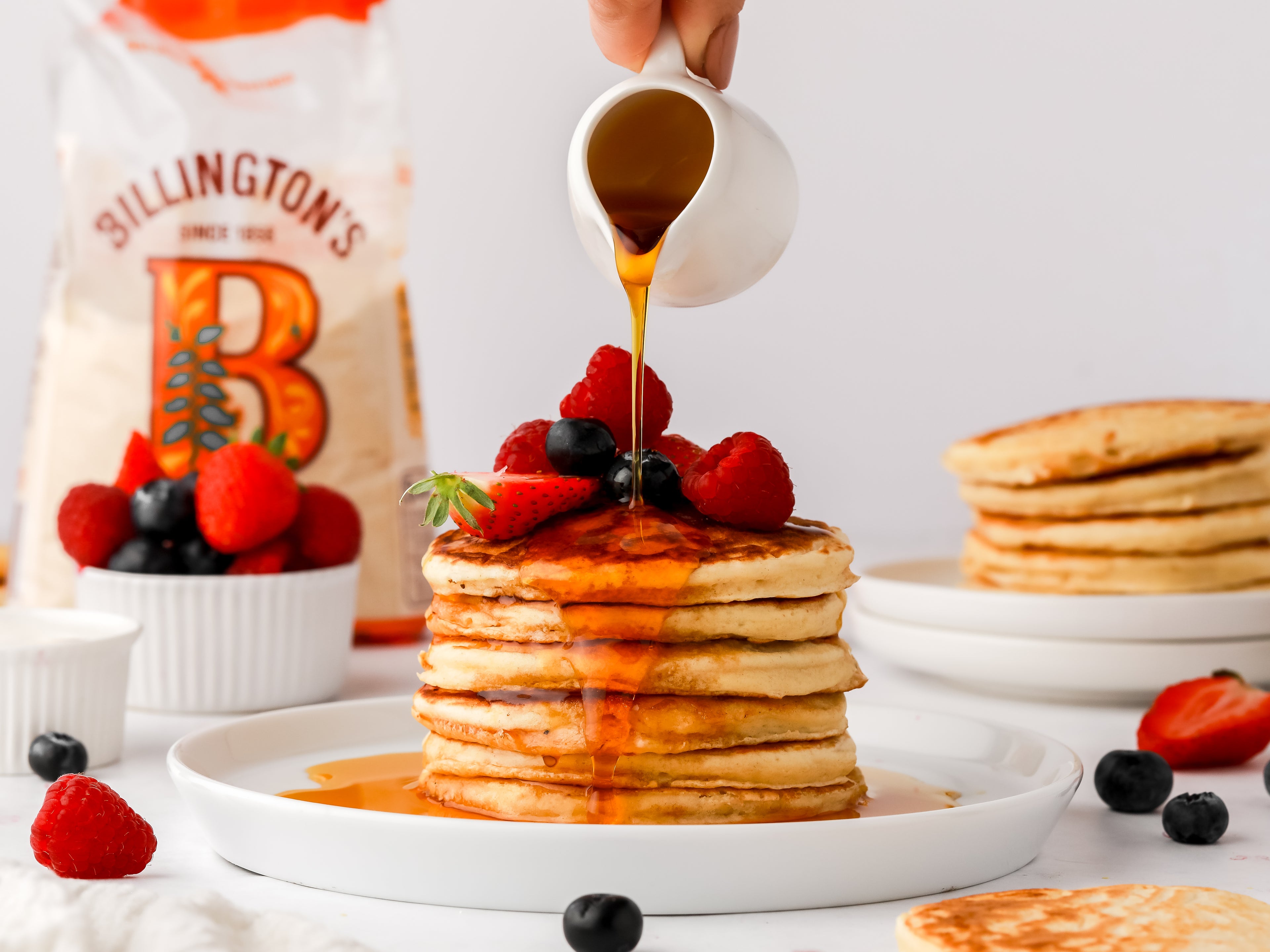 Syrup being poured on top of a stack of pancakes with fresh fruit on