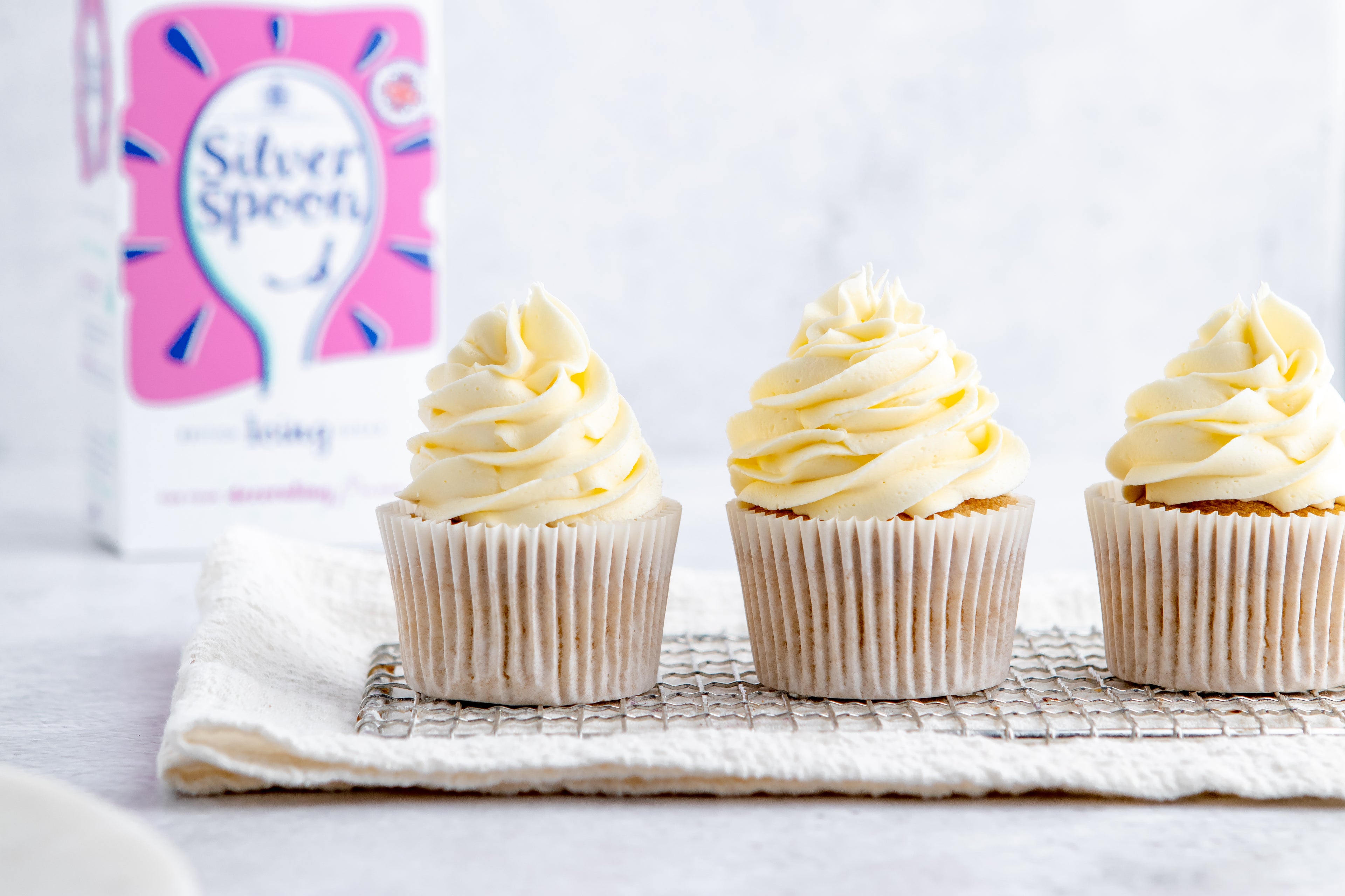 Three Gluten Free Cupcakes lined up on a wire rack, with Silver Spoon Icing sugar in the background