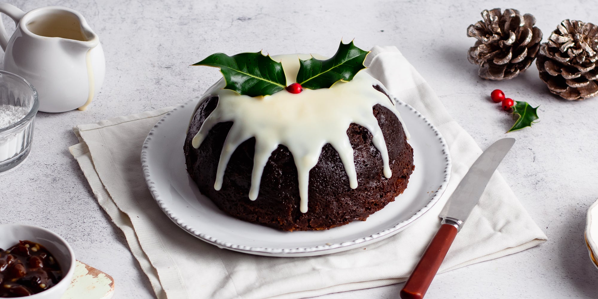 Last Minute Christmas Pudding covered in a generous pouring of cream or brandy butter, dripping down the sides next to a knife and a holly leaf