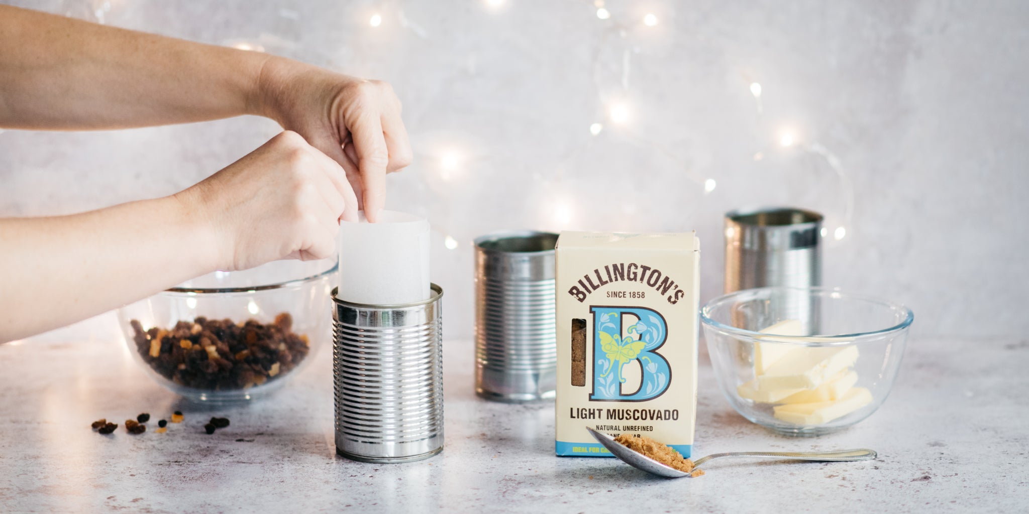 3 tin cans being prepped with baking paper and a bowl of butter, dried fruit and a pack of sugar in shot