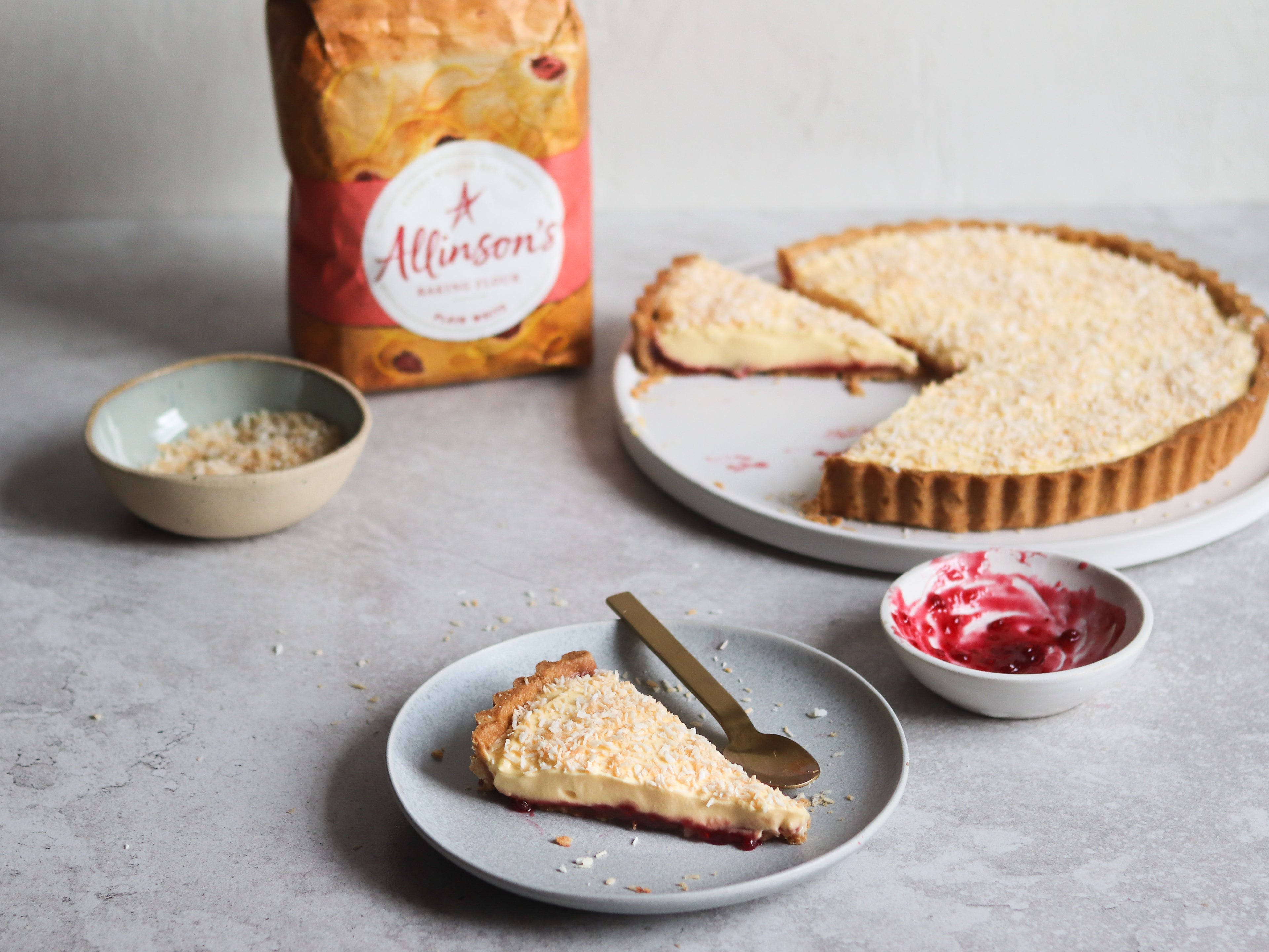 A plate with a slice of Manchester Tart, with a spoon. In the background there is a Manchester Tart with slices cut out of it and a bag of Allinson's Plain Flour