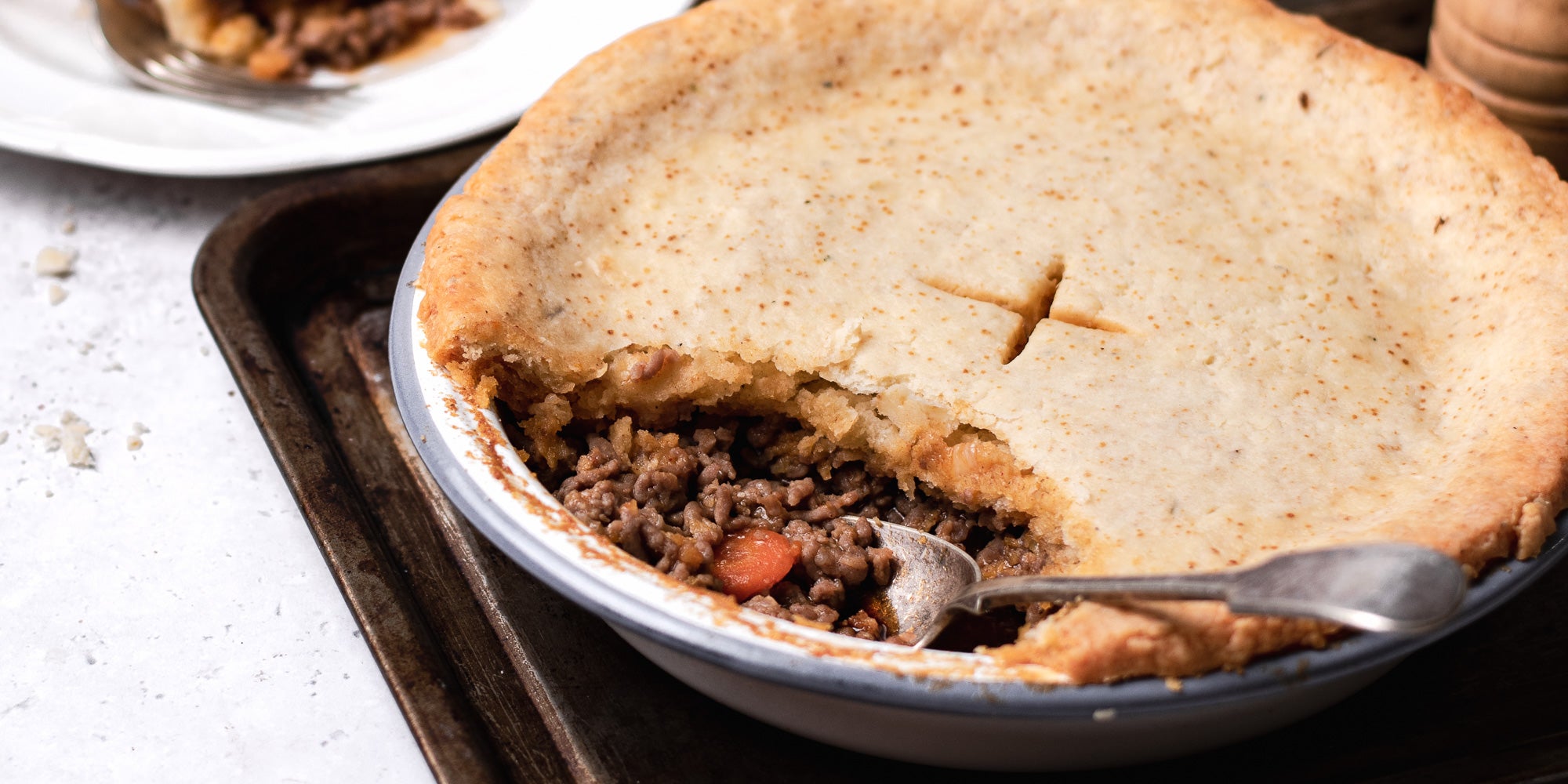 Close up of Suet Crusted Beef & Onion Pie, with the crust open and the filling being scooped out by a serving spoon. Pie is served on a baking tray