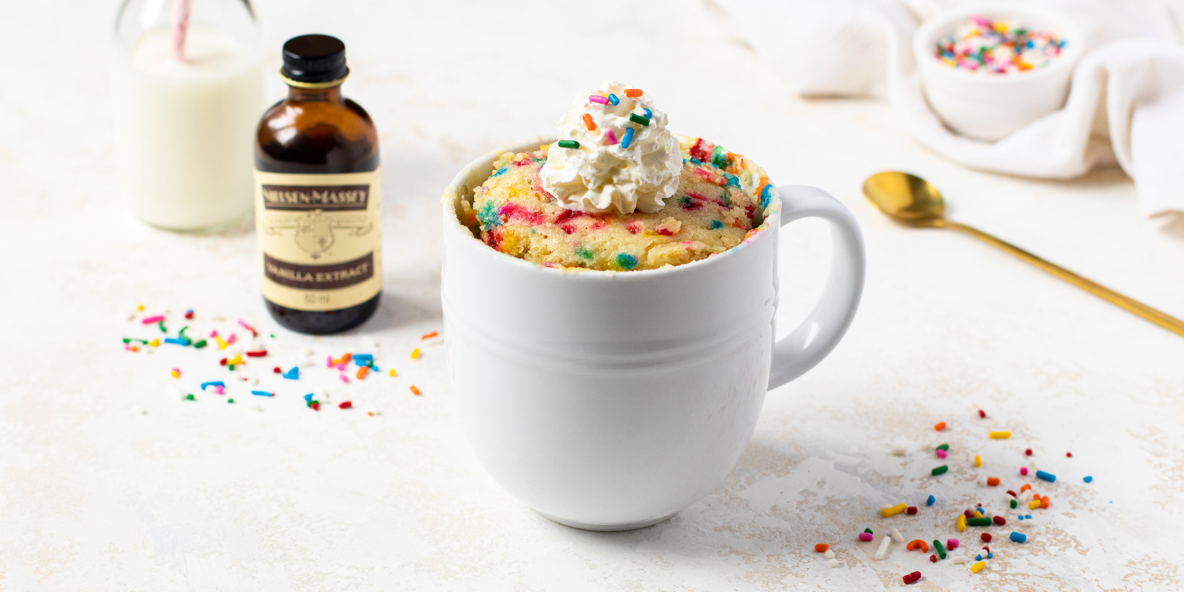 Birthday Cake Mug Cake with a dollop of whipped cream on top with sprinkles, and a bottle of Nielsen-Massey Vanilla extract in the background