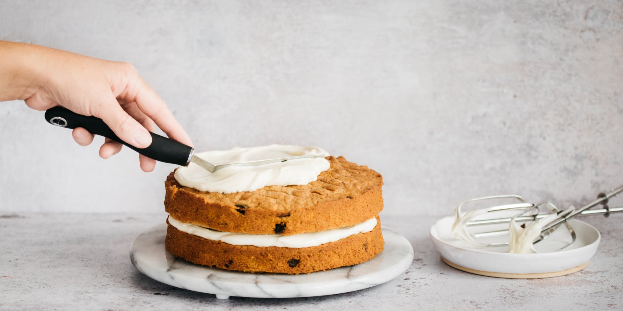 Hand smoothing frosting on top of a cake with a palette knife. Bowl with mixers next to it with icing on it