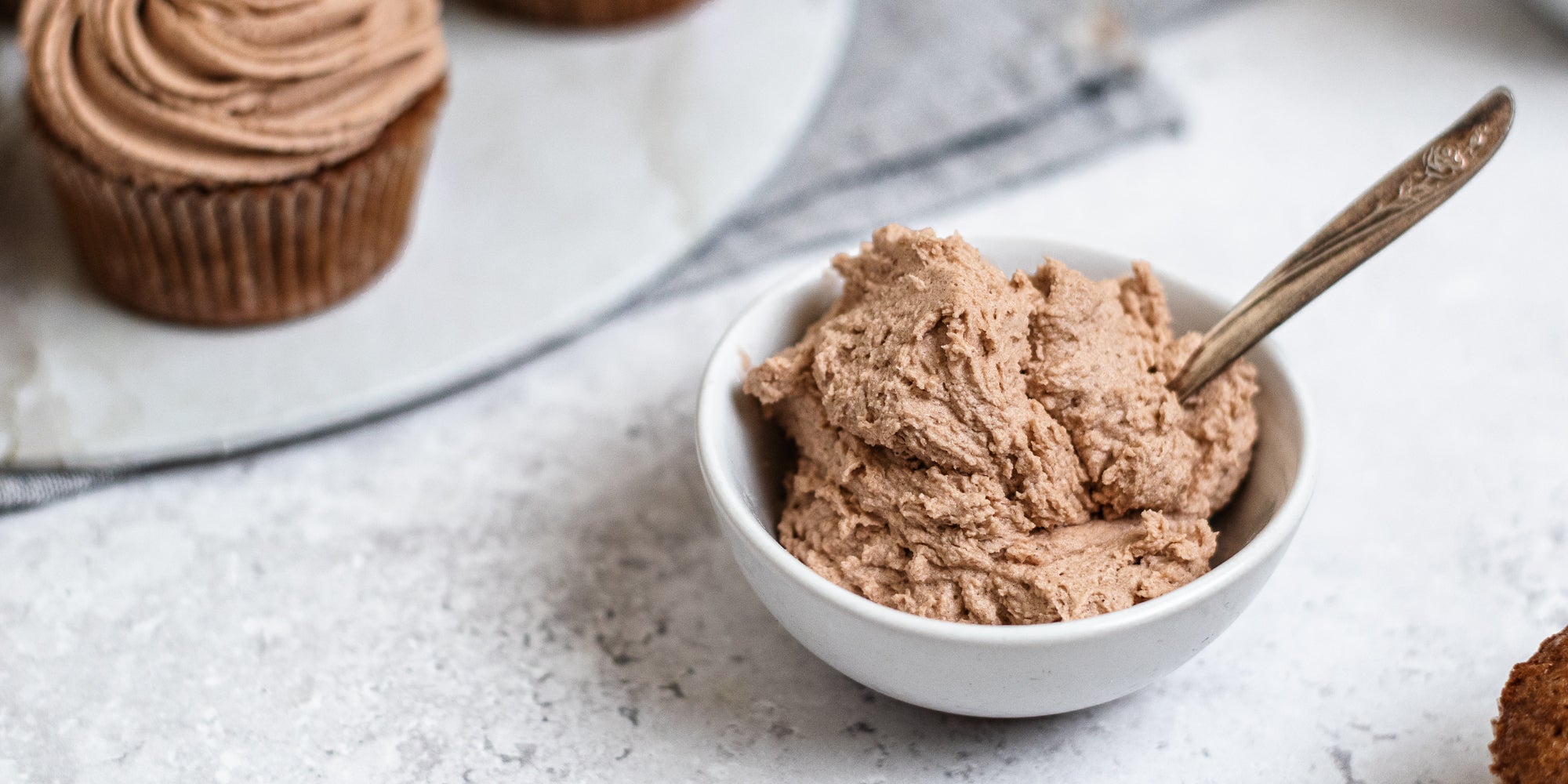 Close up of a small bowl of Dairy Free Vegan Chocolate Buttercream with a spoon dipped in it. Cupcakes topped with Dairy Free Vegan Chocolate Buttercream in the background