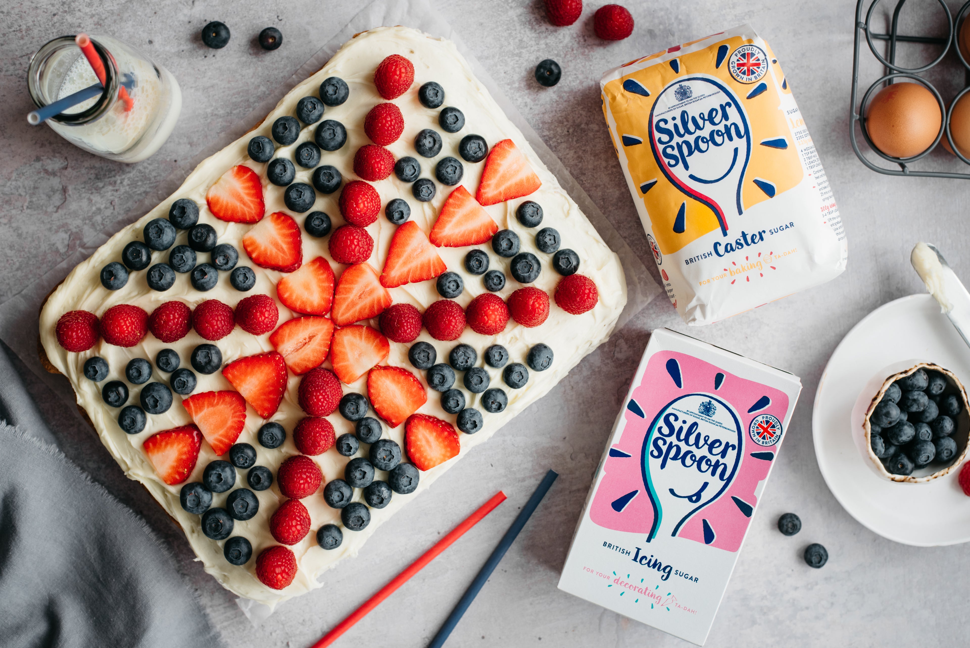 Top view of Union Jack Traybake topped with seasonal fresh berries, next to a bag of Silver Spoon Icing Sugar, and Silver Spoon Caster Sugar