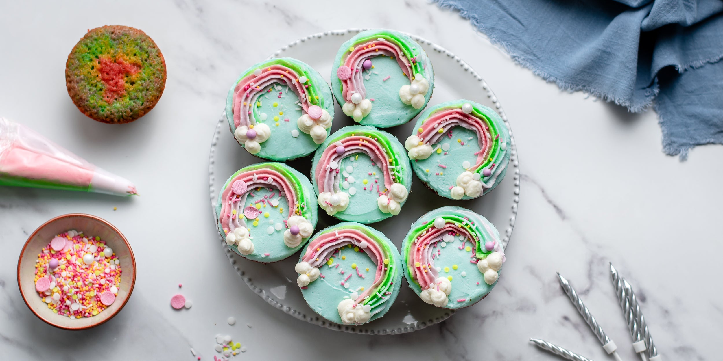 Overhead shot of 7 rainbow decorated cupcakes on a circular plate beside a piping bag, pot of sprinkles and plain cupcakes