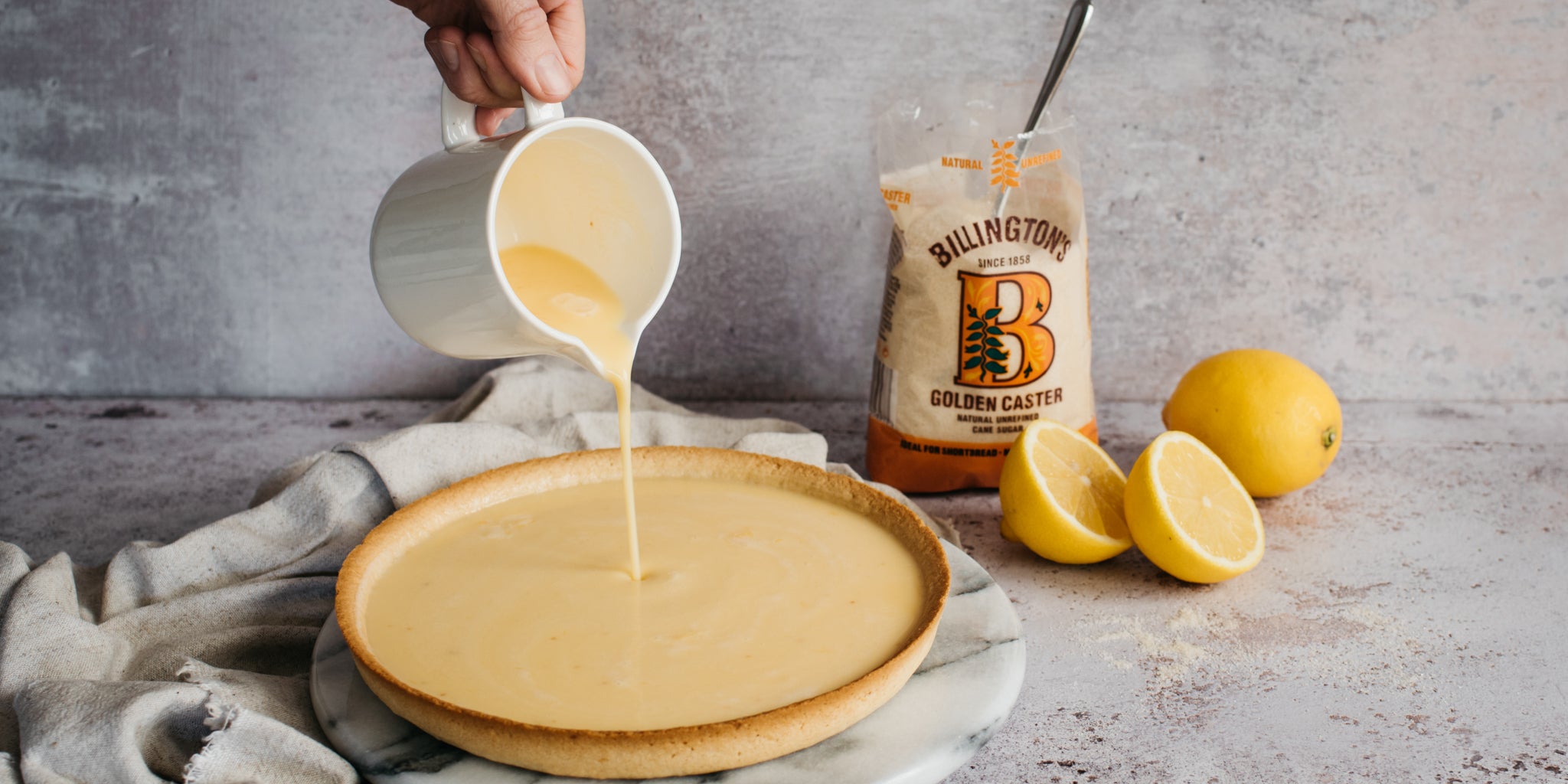 Lemon Tart being filled with hand holding a jug pouring lemon filling into the pie base, next to a bag of Billington's Golden Caster Sugar
