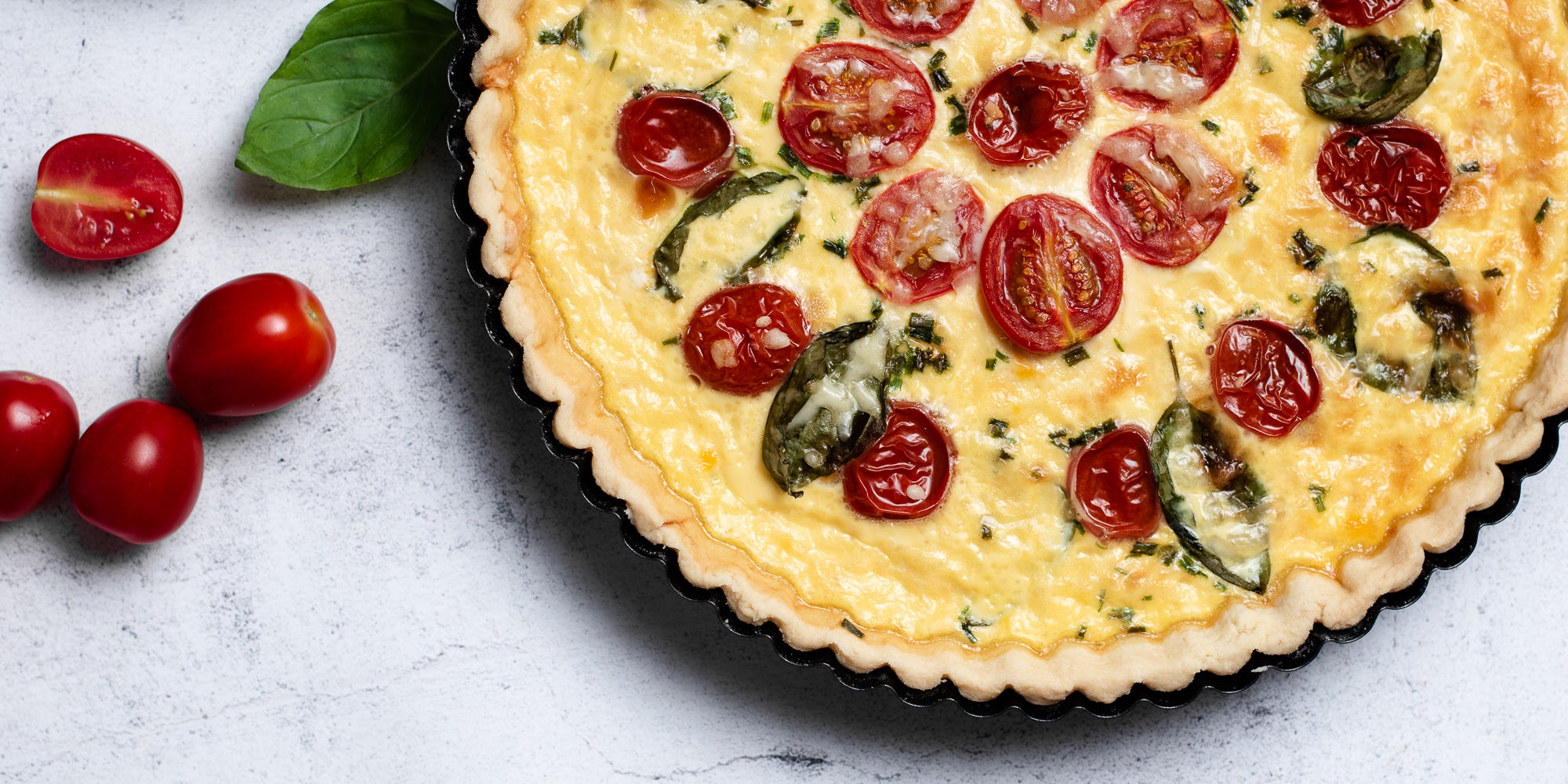 Close up of a Cheese & Tomato Quiche with whole tomatoes and a basil leaf in a pastry baking tray