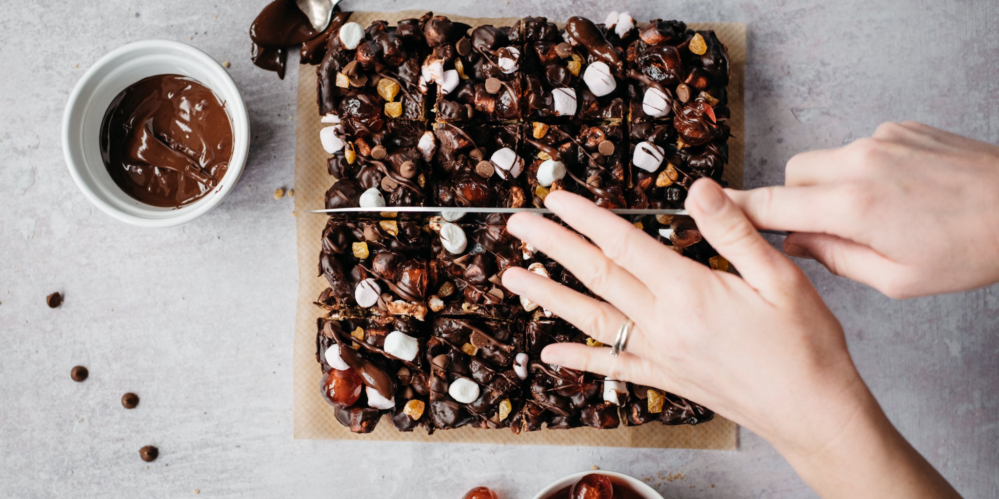 Vegan Rocky Road traybake being sliced into squares with hands holding a knife, next to a bowl of melted chocolate