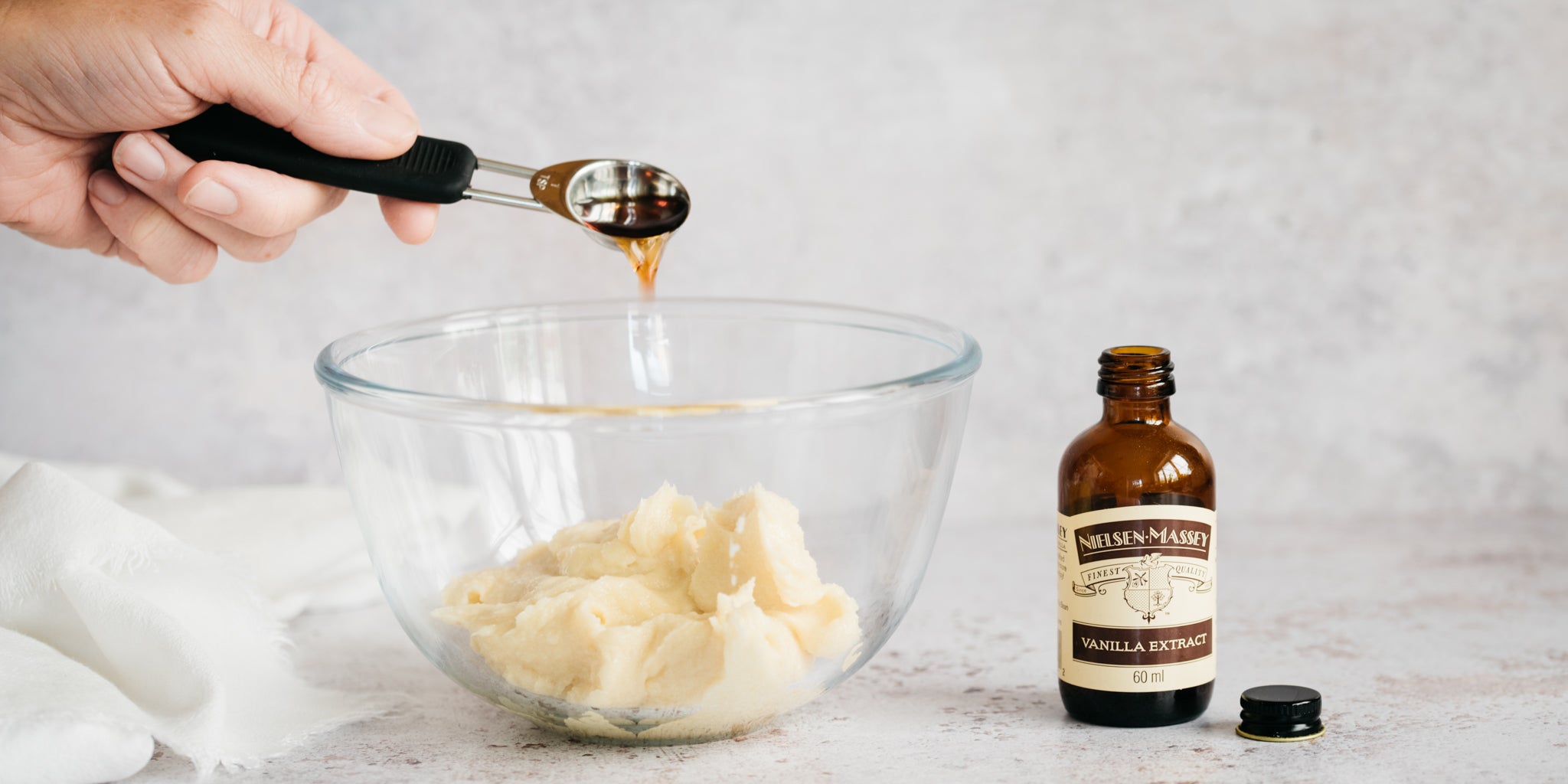 Glass bowl with biscuit mixture in. A hand pouring in vanilla with a measuring spoon. Vanilla bottle next to it