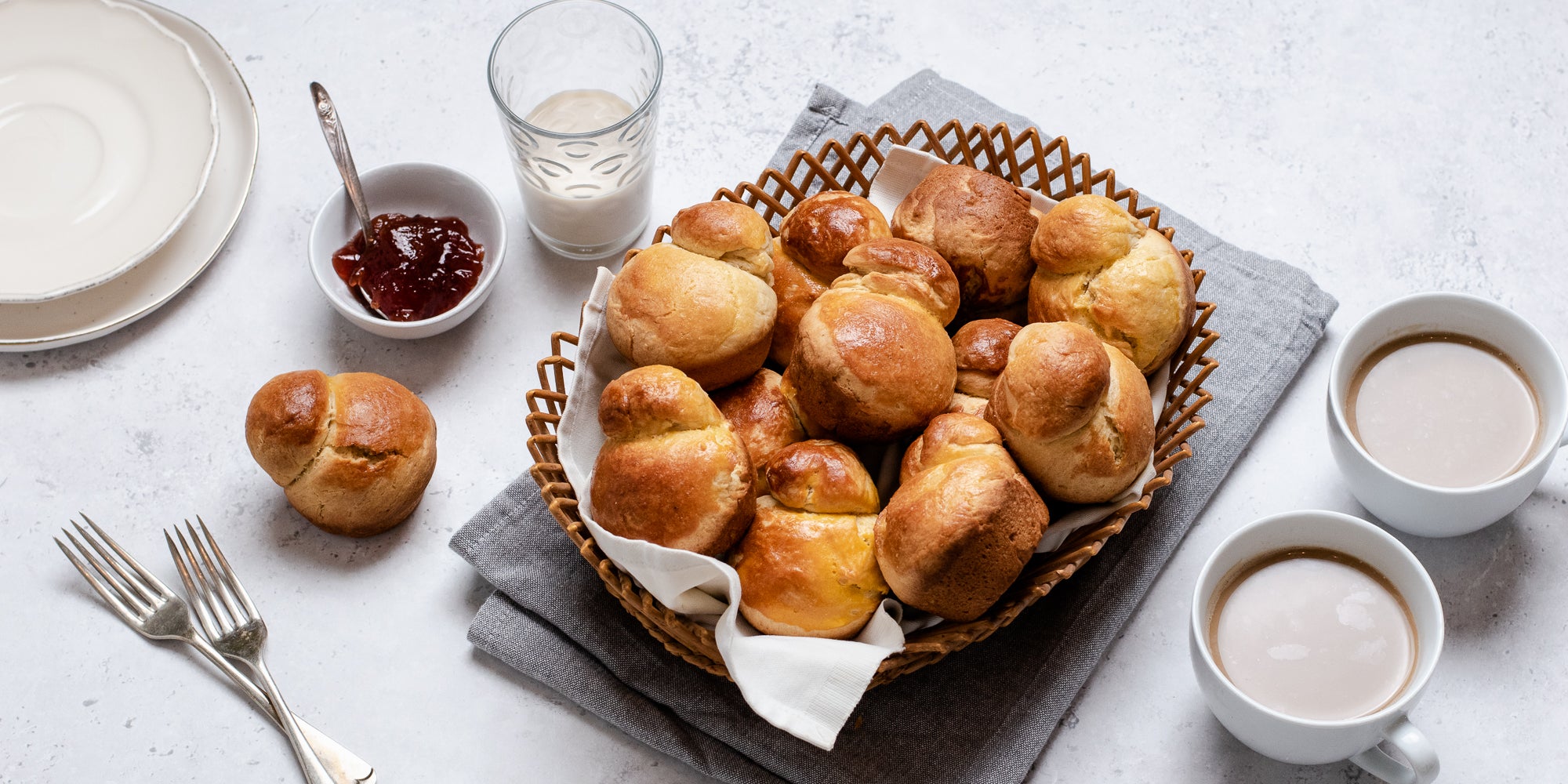 A batch of golden Brioche in a wicker basket, next to cups of tea, a bowl of jam and a glass of milk