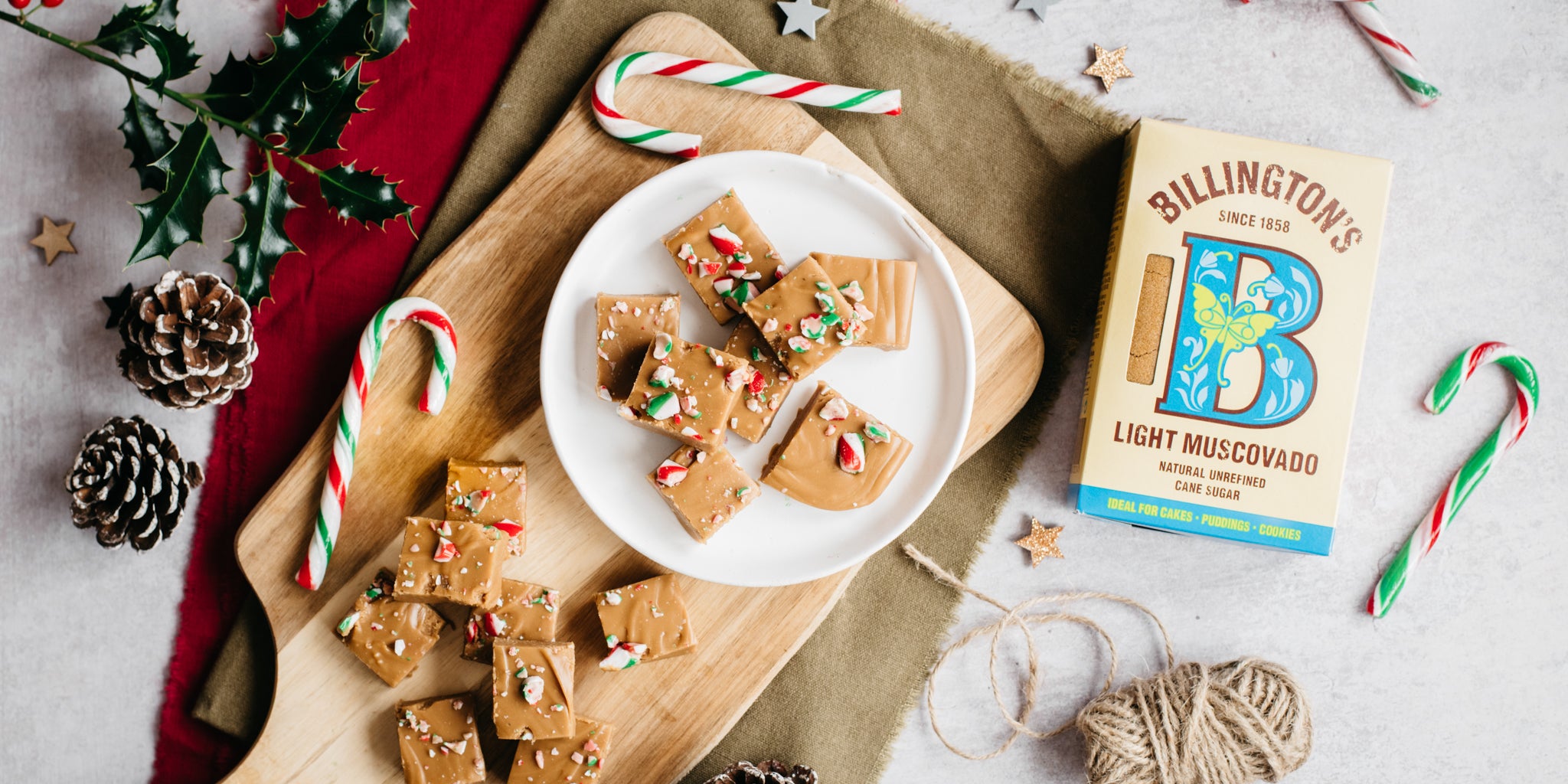 Top view of Billington's Candy Cane Fudge, on a wooden board next to candy canes, pine cones and a box of Billington's Light Muscovado sugar