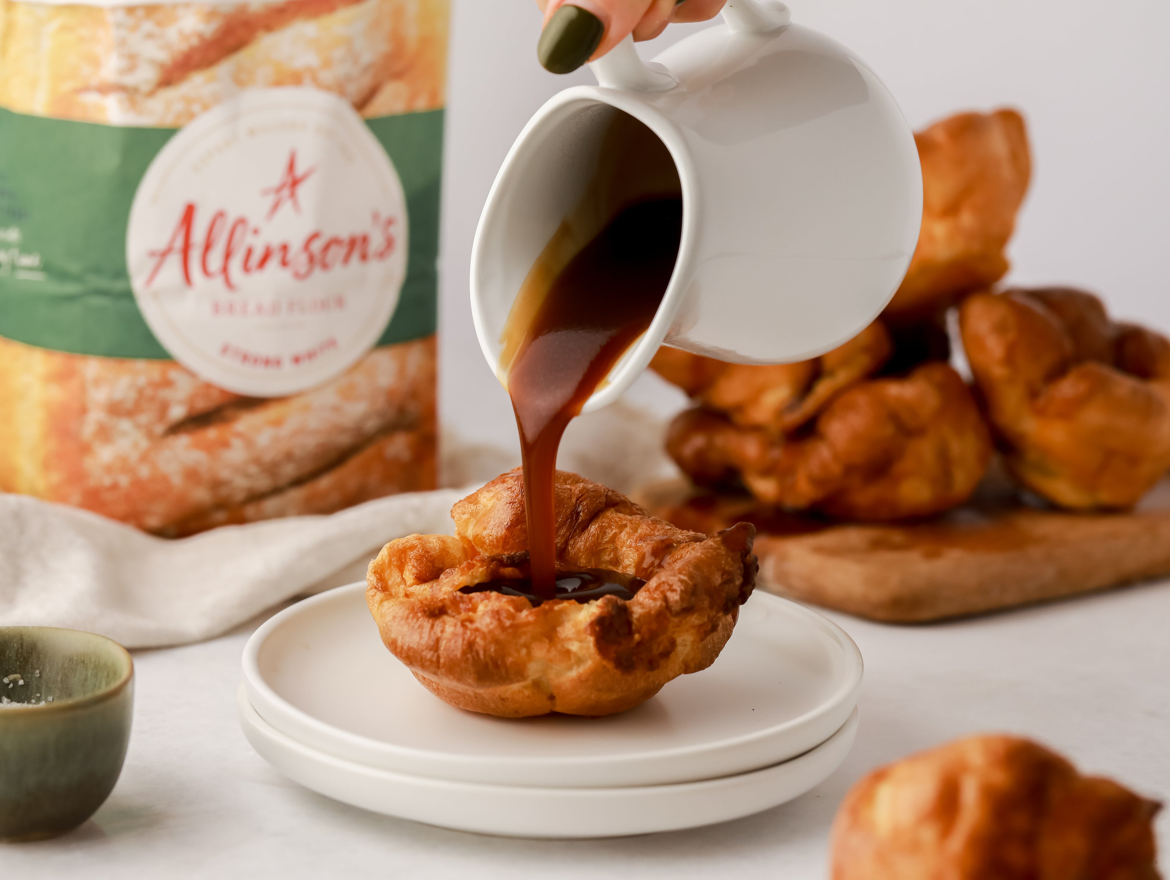 Gravy being poured from a jug into a yorkshire pudding. More yorkshire puddings in the background beside a bag of flour