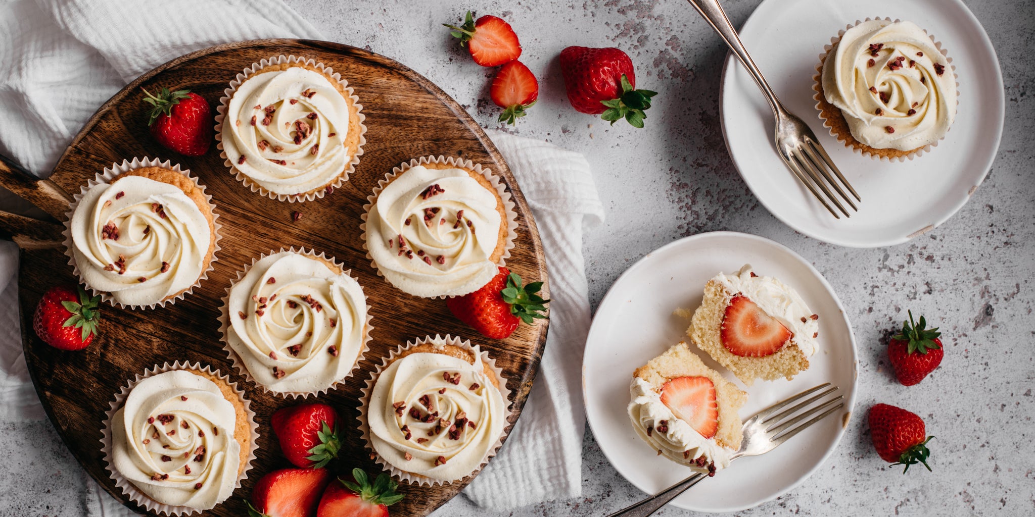 Top view of Strawberry & Prosecco Cupcakes served on a wooden board, with a Strawberry & Prosecco Cupcake cut in half showing the strawberry in the centre of the cupcake