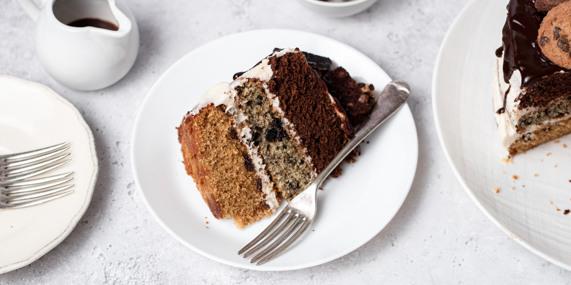 Cookie, Oreo & Brownie Layered Cake slice, on a plate with a fork and a small jug of chocolate ganache