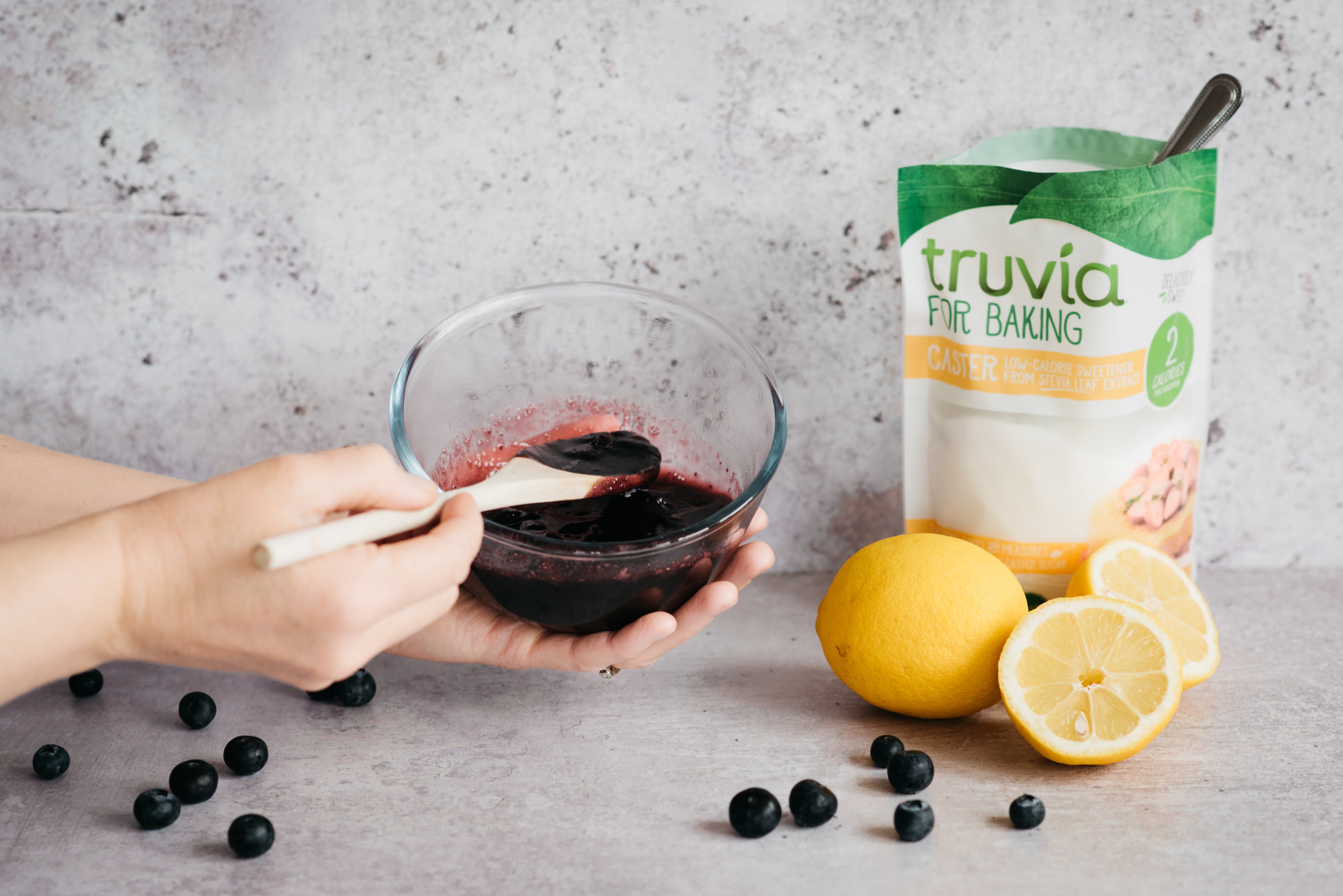 Hand mixing up the blueberry compote next to sliced fresh lemon, and a bag of Truvia for Baking Caster