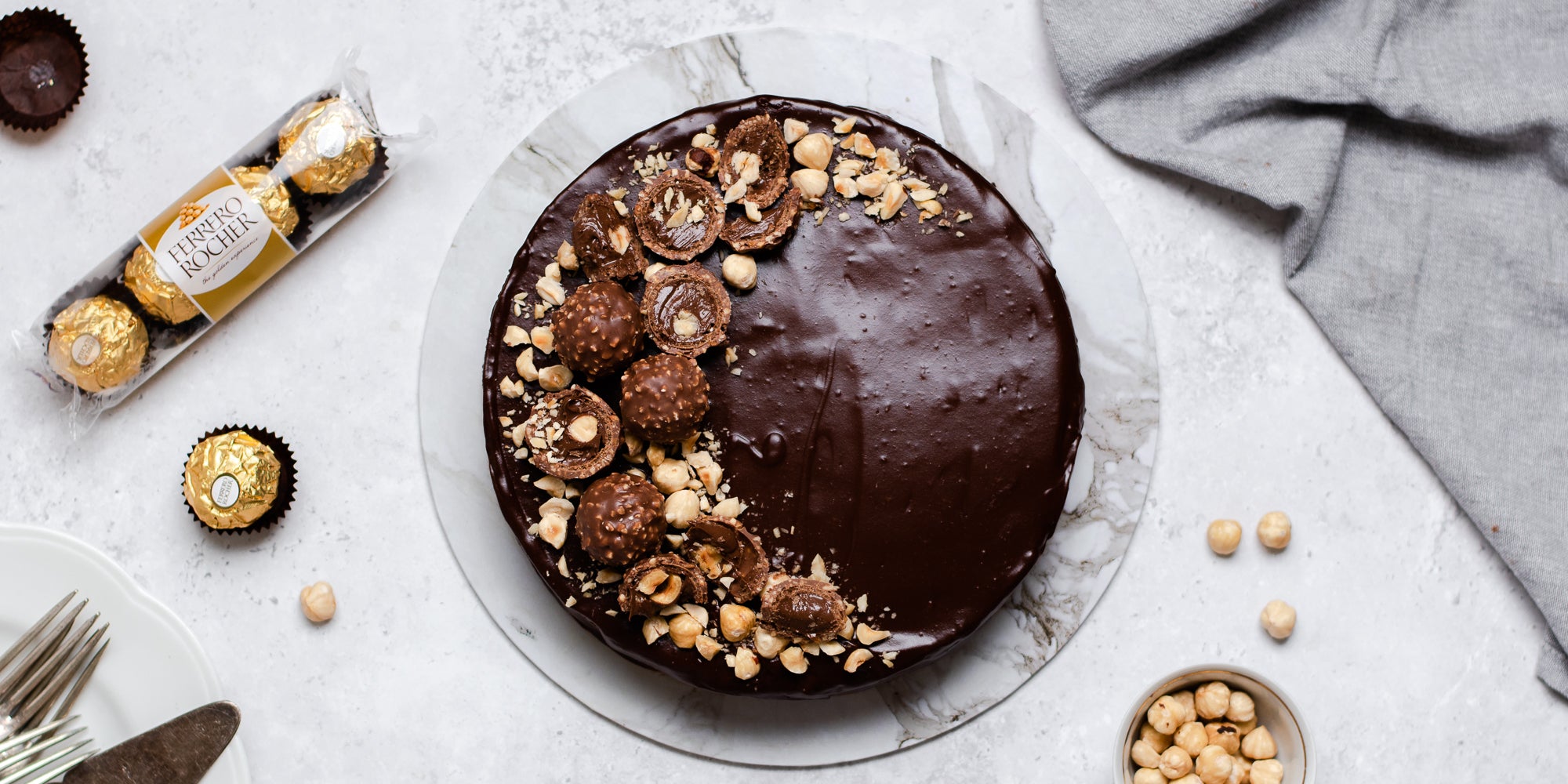 Top view of Gluten Free Chocolate Truffle Sachertorte covered in chocolate icing, topped with chopped Ferrero Rocher's and hazelnuts, next to a packet of Ferrero Rocher's and hazelnuts in a bowl