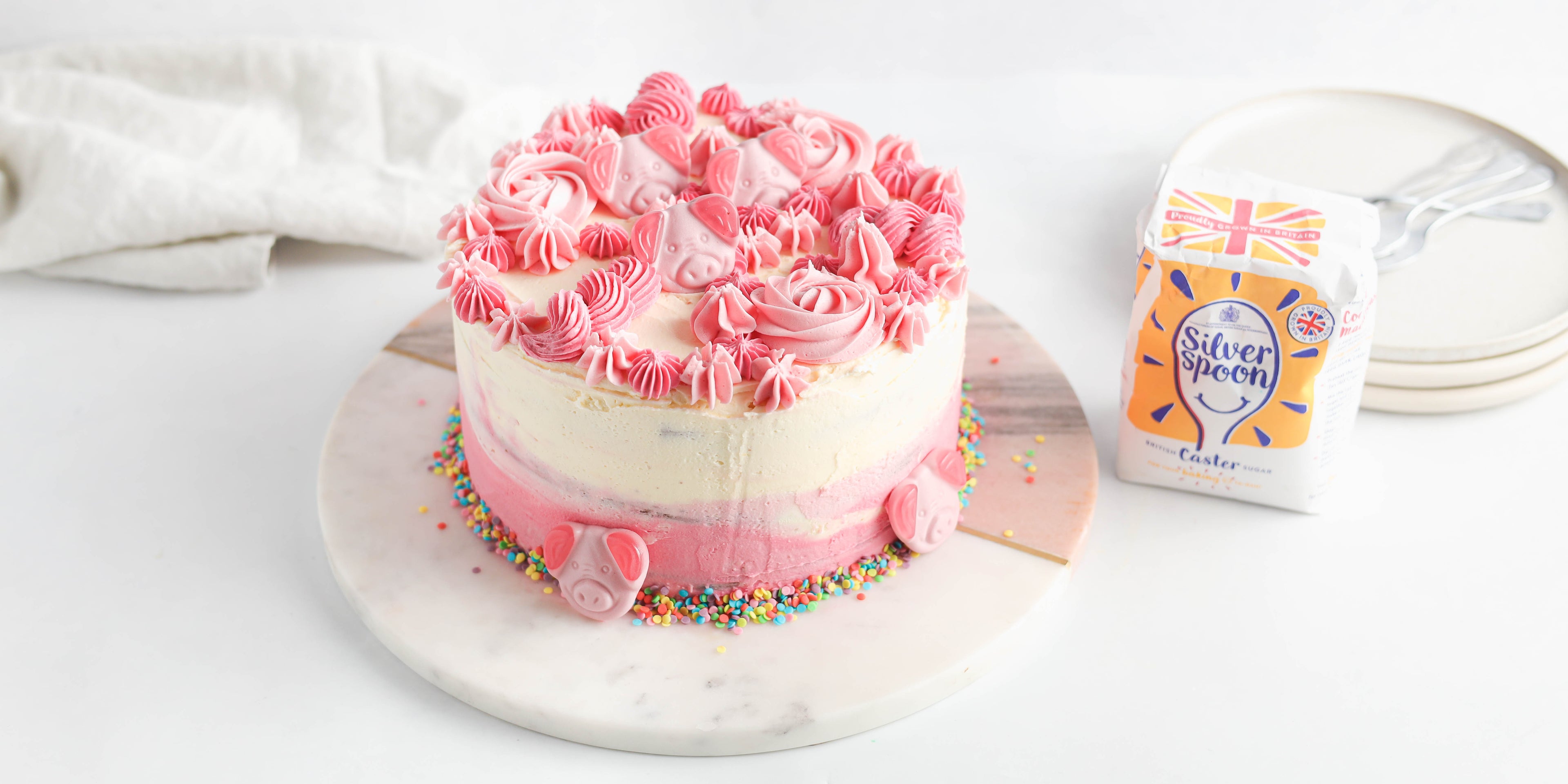Percy Pig Marble Cake decorated with pink buttercream and percy pig sweets, next to a bag of Silver Spoon caster sugar