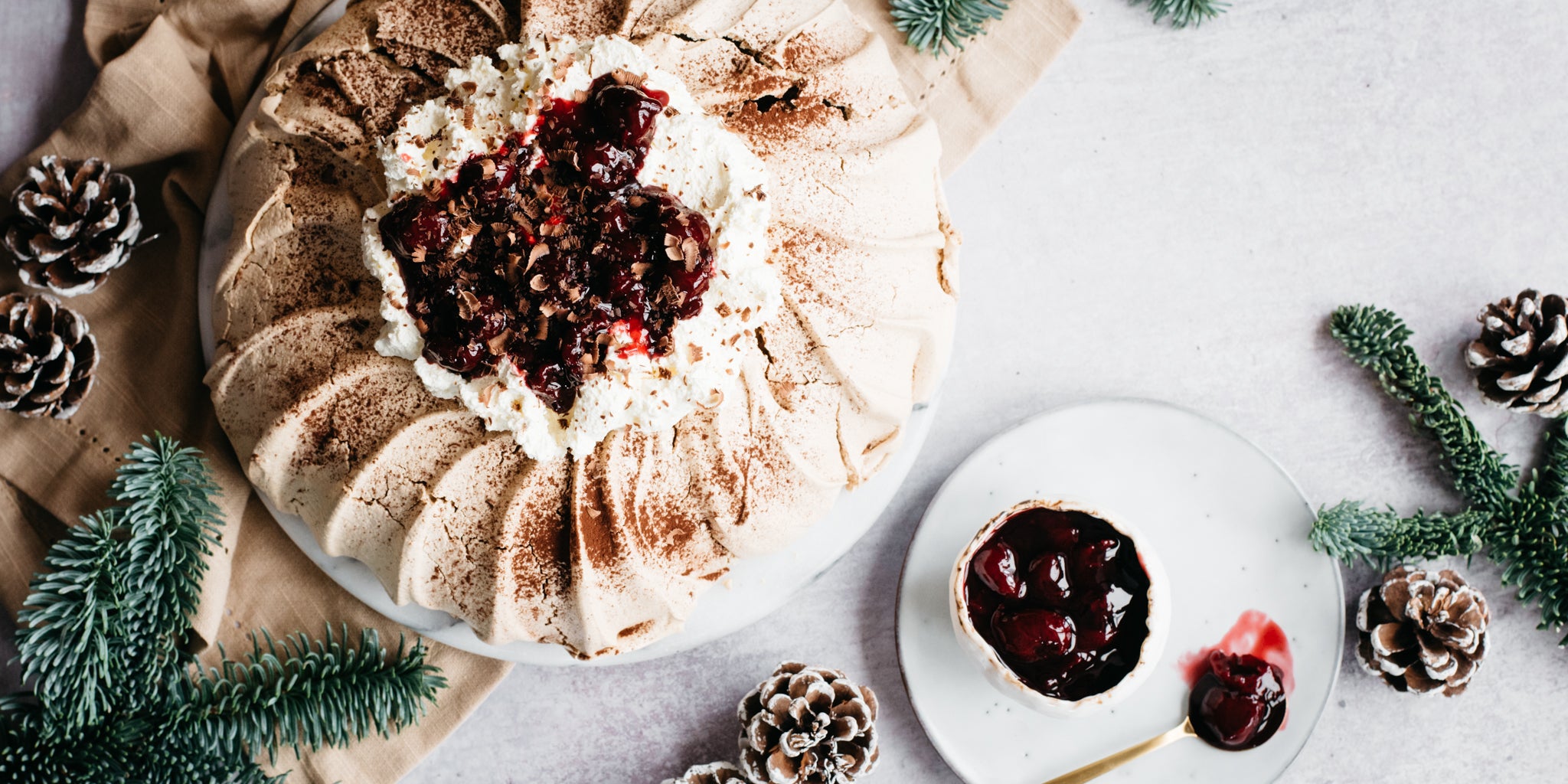 Black Forest Pavlova top view, next to pine cones, leaves and a bowl of cherry kirsch