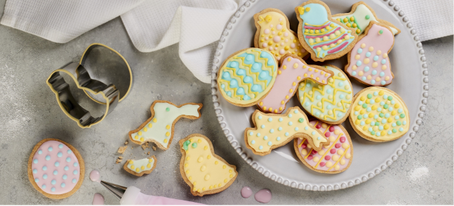 Variety of iced easter biscuits on a white plate beside cookie cutters and a piping bag of pink icing