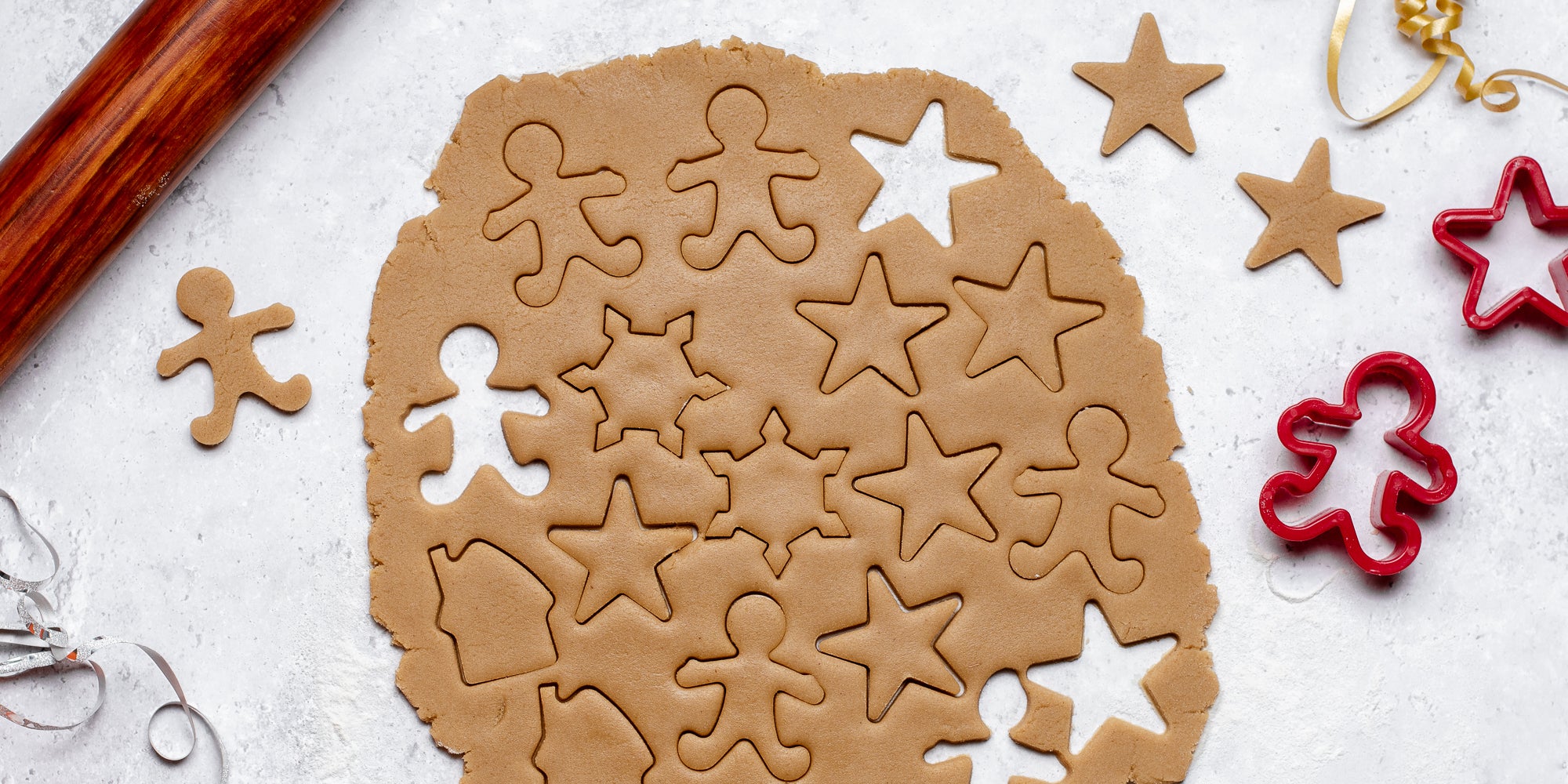 Gingerbread Dough rolled out, with stars, gingerbread houses, and gingerbread men cut out of it, next to a rolling pin and cookie cutters.