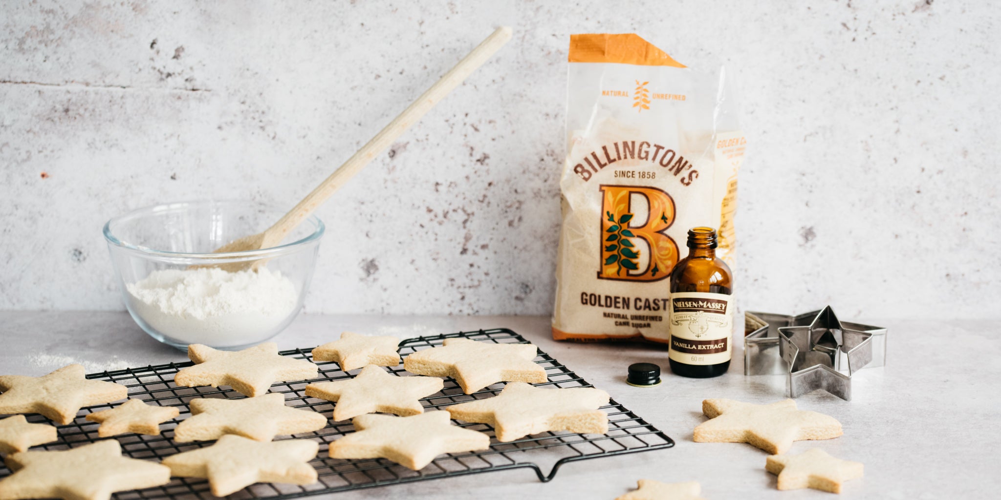 Biscuits on a cooling rack with bottle of vanilla, star cutters, sugar pack and bowl of icing sugar with wooden spoon in